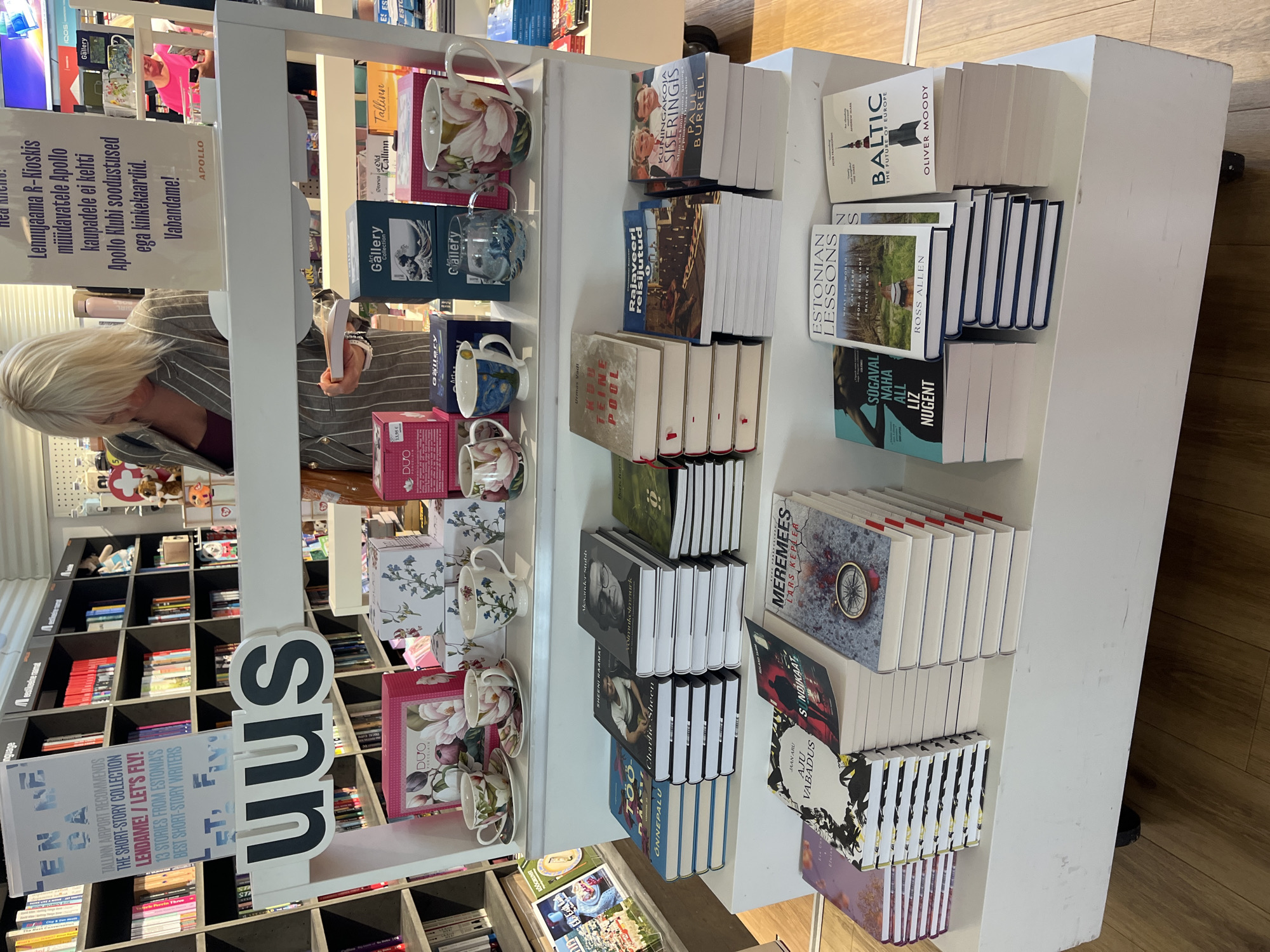 The image depicts a retail display of books and decorative mugs in a store, likely an airport shop in Tallinn, Estonia, given the text on the signage that reads "TALLINN AIRPORT RECOMMENDS" and "LENNAME! / LET'S FLY!". The display is set up on a white, multi-tiered shelf unit. In the background, bookshelves filled with books are visible.
In the foreground, there are stacks of books with prominent titles. On the top shelf, decorative mugs and teacups are arranged. A woman with blonde hair, wearing a striped jacket, is visible in the background, looking down at a book she is holding. The overall lighting suggests an indoor retail environment. The signage also mentions "APOLLO", which is a bookstore chain in Estonia.
