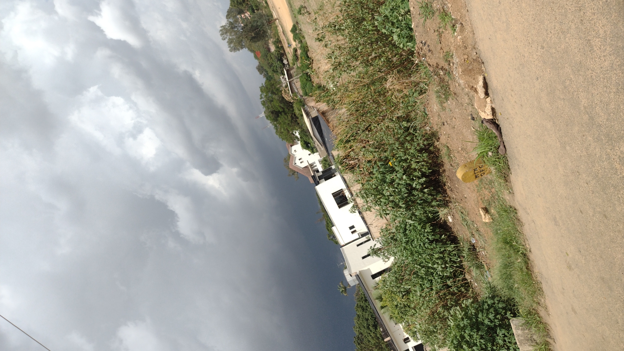 The image captures a section of Jos, Nigeria. The frame is angled, presenting a view of the sky dominated by dark storm clouds on the left side. Adjacent to the sky is a cluster of buildings. Beyond the buildings, the scene transitions into a roadside environment. The right side features vegetation growing along the roadside, with a yellow milestone marker visible near the edge of the paved road.