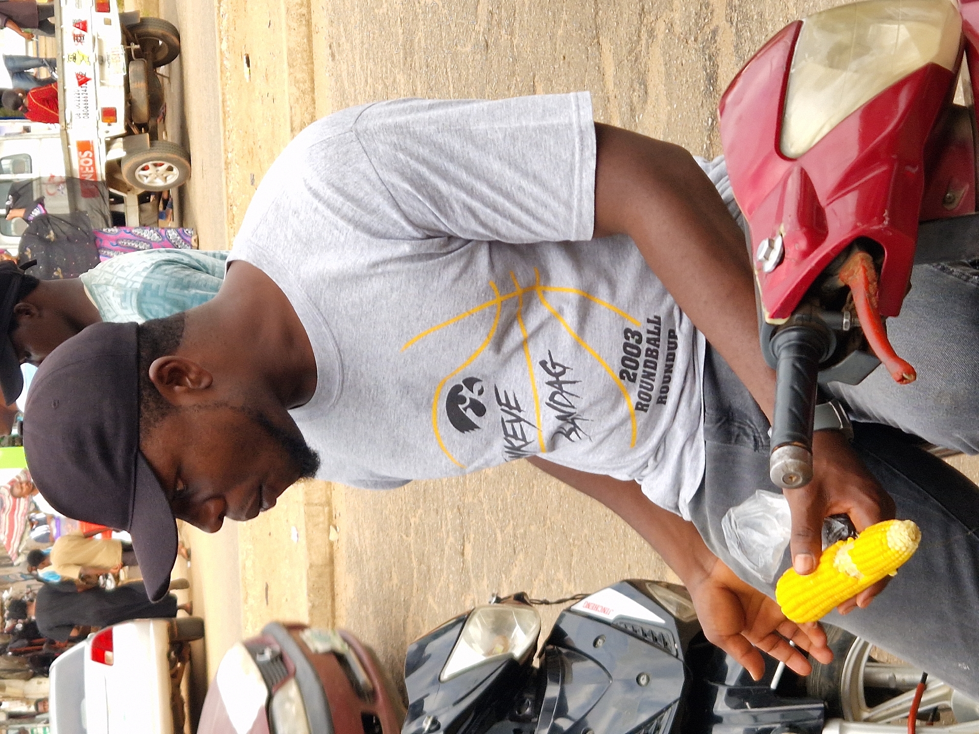 A man wearing a black baseball cap and a grey t-shirt is seated on a red motorcycle, holding a partially eaten ear of corn. He is looking to his right, with his left hand extended and holding the corn. The man is wearing dark grey jeans. In the background, a white pickup truck with a white cargo bed is visible, parked on a dirt road. Several other people are blurred in the background, some walking and others standing near the truck. There are also a few cars and other motorcycles visible in the distance. The environment appears to be an outdoor market or street scene, with a dusty ground and what looks like buildings in the far background.