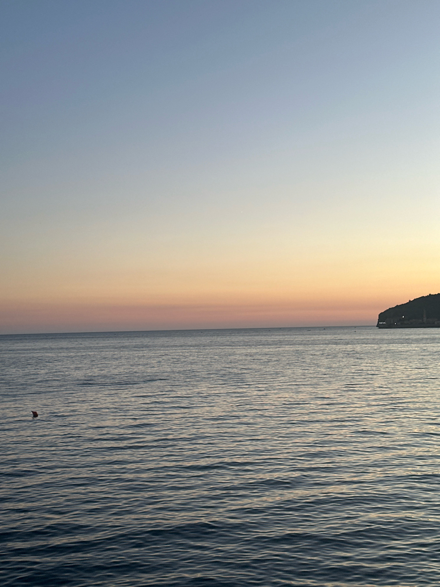 The image depicts a serene seascape at dusk, likely near Budva, Montenegro. The horizon is painted with soft hues of blue, transitioning through pale pink and orange, indicating the twilight hours. The sea is calm, with gentle ripples reflecting the fading light. In the distance, a dark, hilly coastline is visible, dotted with lights that suggest human habitation. The coastline appears to be a headland, possibly forming part of Budva's coastal landscape, known for its rocky promontories and picturesque settlements. A small red buoy floats on the left side of the water, adding a subtle point of interest to the expansive ocean. The overall atmosphere is tranquil and visually appealing, capturing the quiet beauty of a coastal evening.