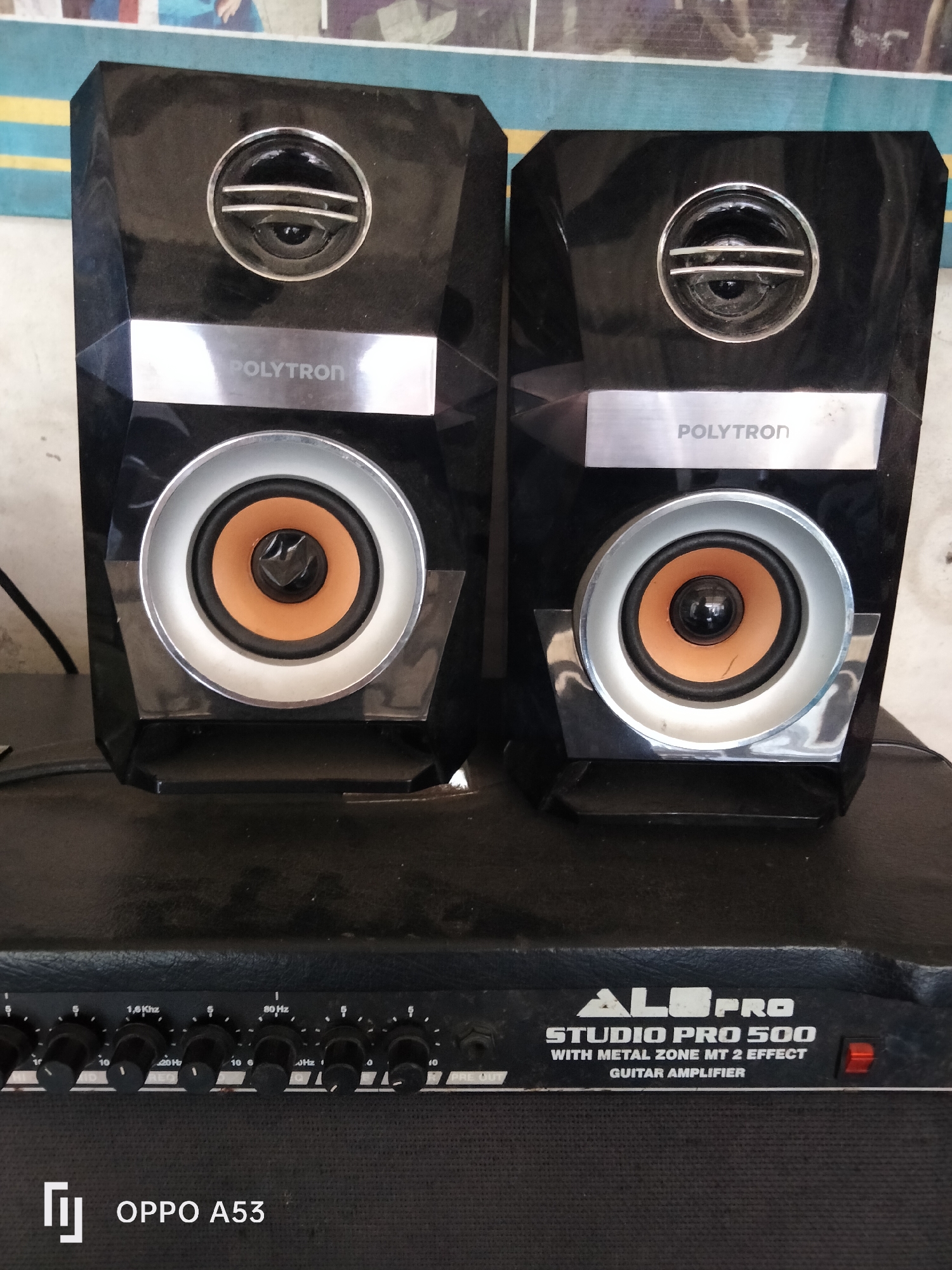 The image shows a pair of black POLYTRON speakers positioned in front of a black GUITAR AMPLIFIER. The amplifier has various knobs and labels, including "ALOPRO STUDIO PRO 500 WITH METAL ZONE MT 2 EFFECT GUITAR AMPLIFIER." The speakers are tall and angular, with silver accents around the circular speaker cones and a silver strip with the POLYTRON logo above each speaker. The amplifier is resting on a dark surface, and the speakers are placed on top of it. The overall setting appears to be indoors, with a blurred background that suggests a wall or backdrop. The lighting is adequate, highlighting the details of the electronic equipment. The image does not contain any people or specific urban landmarks that would indicate a precise location within Denpasar, Indonesia.