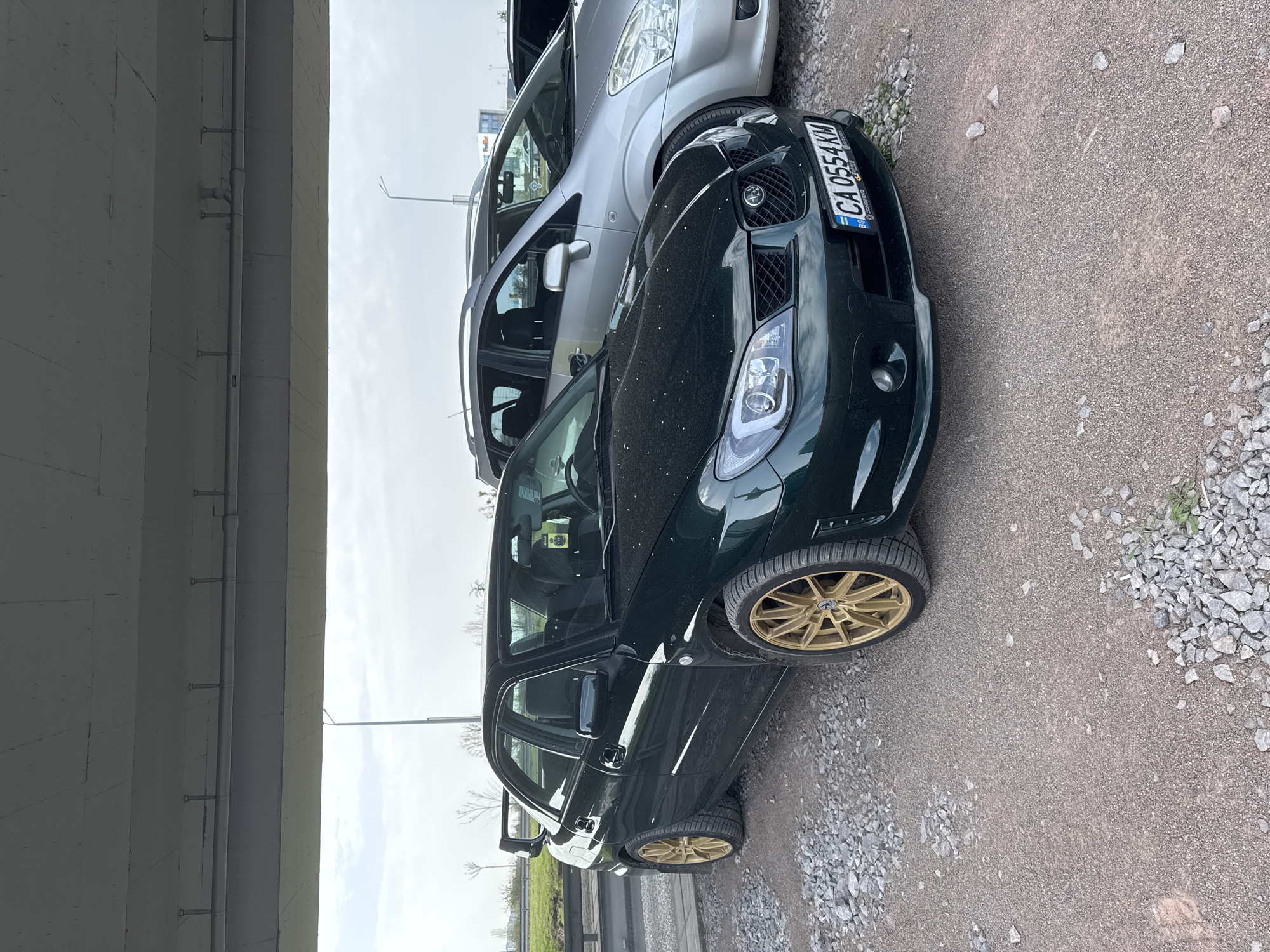 The image shows a dark green Subaru Impreza parked on a gravel surface, under what appears to be a highway overpass. The car has gold rims and a Bulgarian license plate that reads "CA 0554 KM". Next to it is a silver-colored car, partially visible. The background includes the concrete structure of the overpass, a roadside barrier, and some greenery. The location is likely within the city of Sofia, Bulgaria, based on the license plate. The lighting suggests an overcast day.
