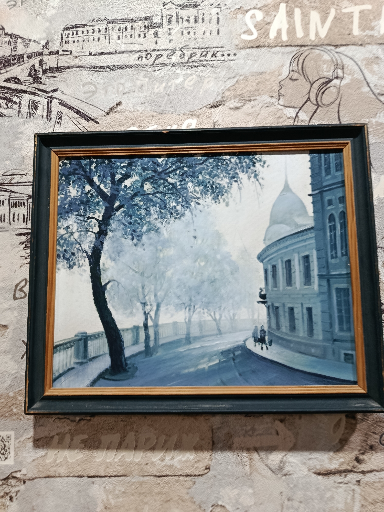 A framed oil painting depicting a street scene in muted blue tones is hung on a textured wallpapered wall. The painting shows a curved street lined with trees and a low wall. On the right side of the street, there is a blue building with a dome-like structure and several windows. Two indistinct figures are walking on the street. The wallpaper behind the painting features a sketch of a city skyline on the left and a profile of a woman with headphones on the right, with handwritten text in Russian. The overall impression is a cool, atmospheric urban landscape. The provided image does not contain any specific elements that would allow for the identification of the location as Apatity, Russia.