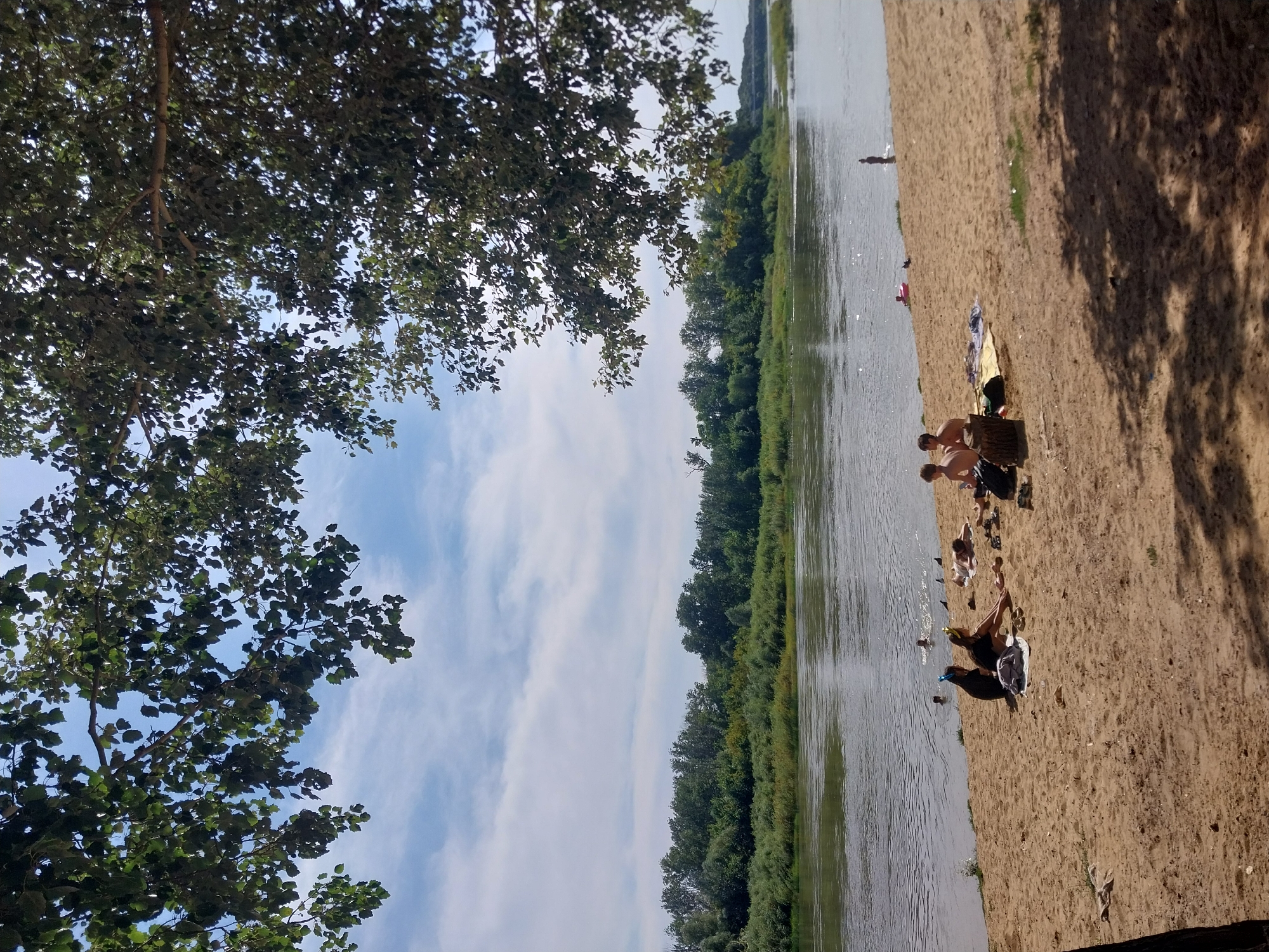 This image depicts a sunny day on a sandy riverbank, likely in Orenburg, Russia. The foreground is dominated by a light brown sand beach, with dappled shadows from overhead trees creating a textured surface. Several people are visible on the beach. Two young individuals, appearing to be male, are sitting on the sand. Further down the beach, two individuals, seemingly female, are also sitting. To the left, several other people are in the water, some swimming and others standing. A pink inflatable ring is visible in the water. The river itself is wide and appears calm, with its surface reflecting the sky. Across the river, a dense line of green trees forms the opposite bank, with a hazy, distant shoreline in the background. The sky is blue with scattered white clouds. The overall scene suggests a relaxed, recreational atmosphere on a warm day by the water.