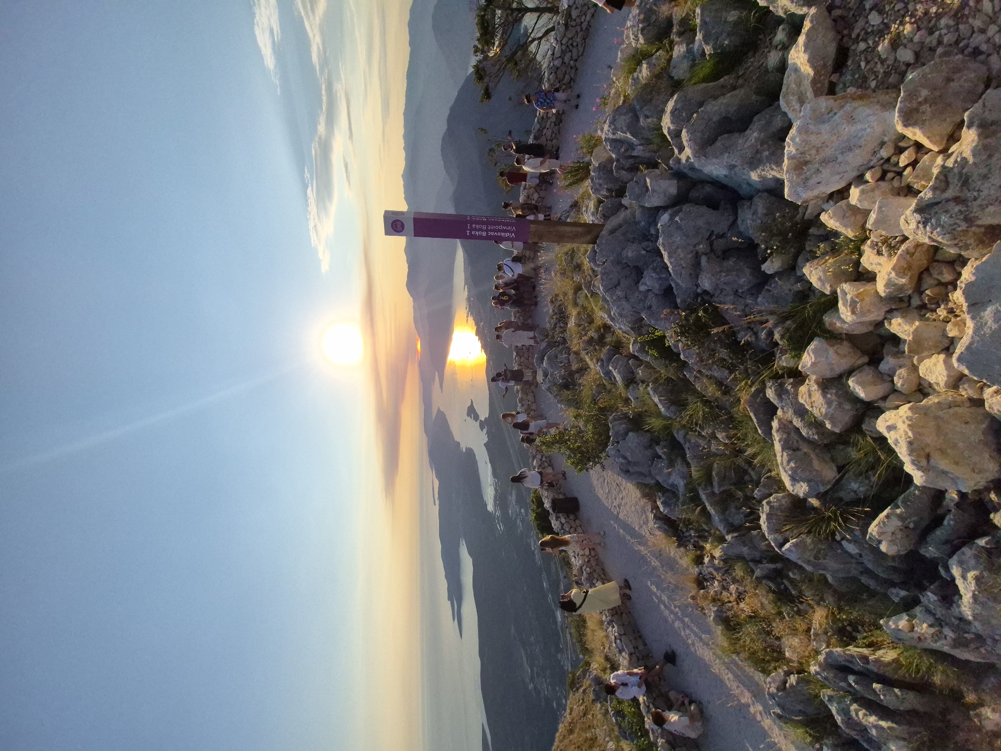 The image captures a panoramic view from a rocky, elevated vantage point overlooking a body of water and distant mountains at sunset. The sun is low on the horizon, casting a golden, shimmering reflection on the water and creating a warm, orange glow in the sky with some wispy clouds. A group of people are gathered on a paved path, some standing and observing the view, others sitting or taking photos. A purple and white signpost with text is visible in the foreground, indicating a viewpoint. The terrain is rugged, with large rocks and sparse vegetation. The overall scene depicts a popular tourist or scenic spot, likely a viewpoint in a coastal or mountainous region. The location is identified as Vidikovac Bol, Viewpoint Bol 1, on Vidikovac, in Kuk, Montenegro.