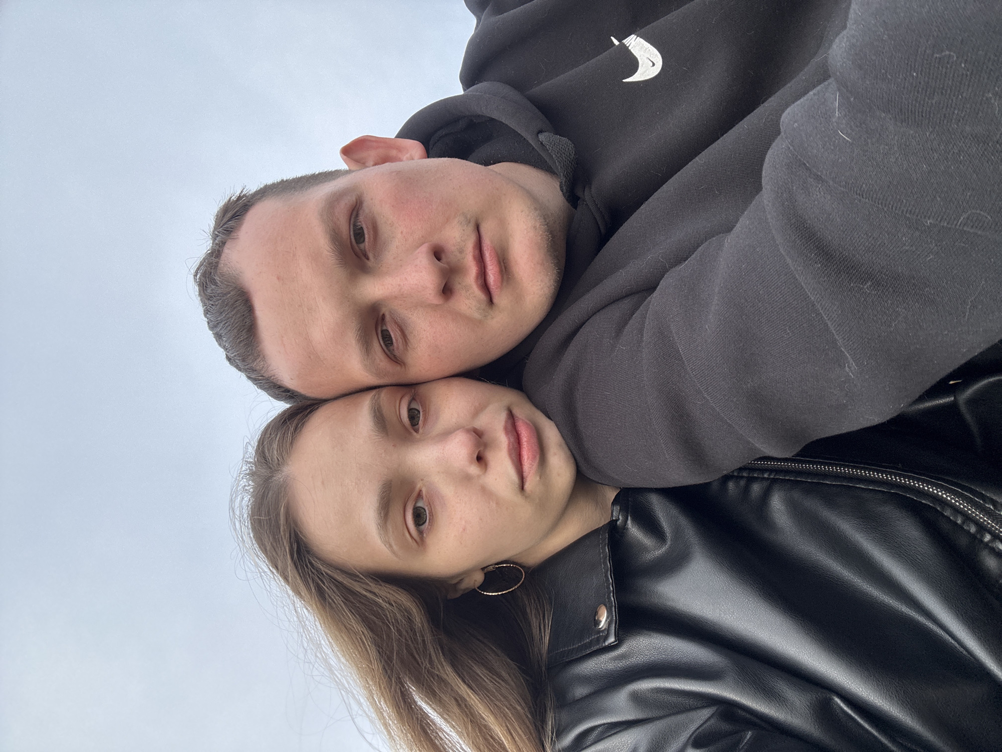 A close-up, low-angle selfie captures two individuals, a man and a woman. The man, positioned above, wears a black hooded sweatshirt featuring a white logo. His face is visible, revealing his features. The woman lies beneath him, wearing a black leather jacket and simple golden hoop earrings. Their faces are close together, both looking towards the camera. The background shows a cloudy sky, indicating an outdoor setting. The specific location in the city of Olkhovka, Russia is not evident from the provided image.