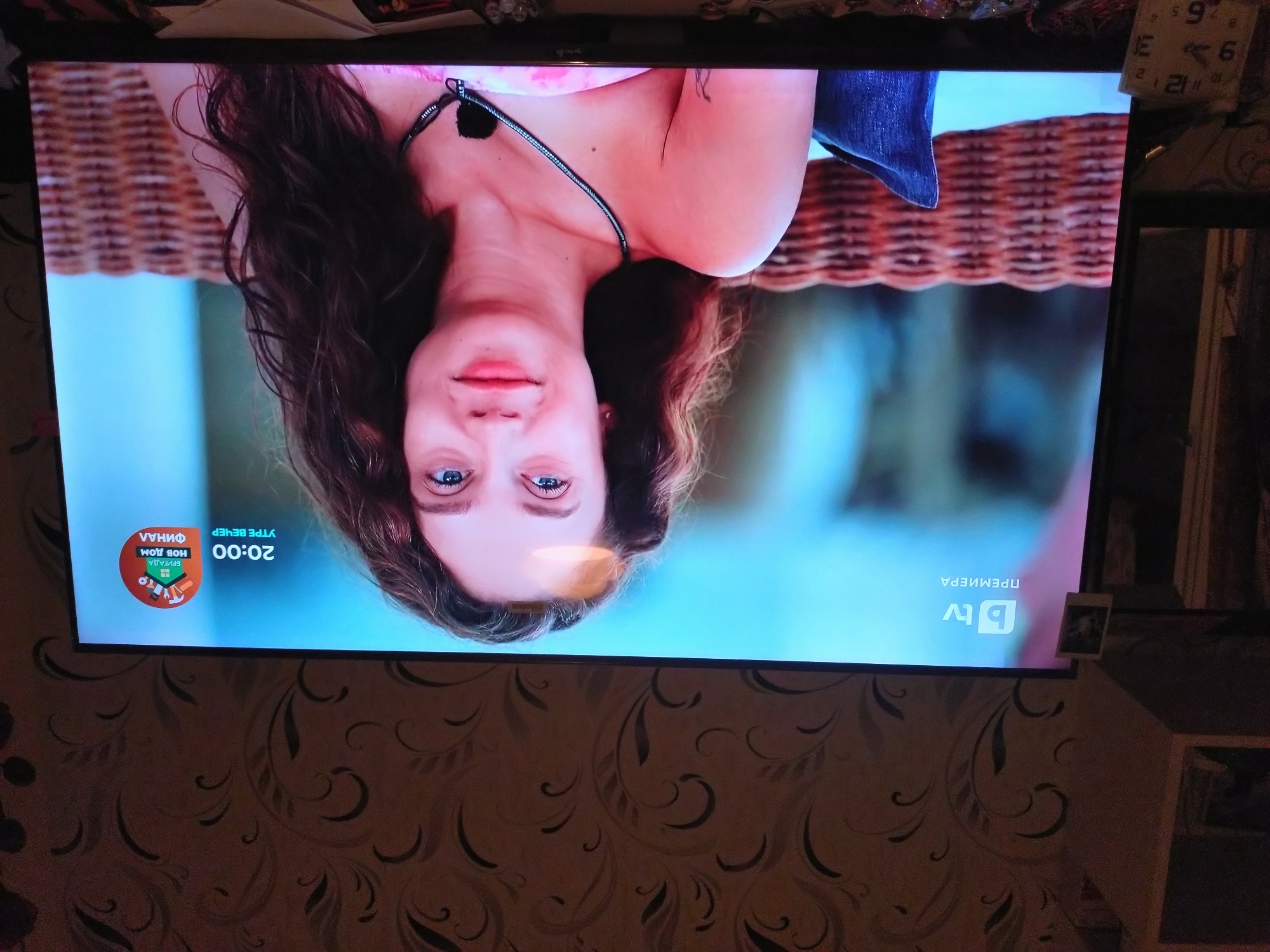 A woman is seen on a television screen. Her head is oriented downwards. She has long, dark hair, fair skin, and blue eyes. She is wearing a top with thin straps. Part of a denim garment is visible near her shoulder. In the corner of the TV screen are logos and text in Bulgarian, including "bTV" and "20:00." The television is mounted on a wall covered in patterned wallpaper. A clock and other items are partially visible on top of the television. A shelf is to the right of the TV. It is not possible to determine the location in Stara Zagora, Bulgaria based on the image.