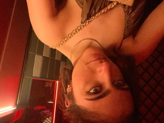 A medium-close-up selfie captures a young adult female from the chest up, positioned slightly off-center to the left. She has dark hair, brown eyes, and a neutral expression, looking directly at the camera. She wears a light brown or olive-colored top with thin straps, a delicate gold-colored chain necklace, and a gold-toned stud earring in her visible right ear. A gold-colored chain strap from a bag or purse is draped across her right shoulder.
The indoor background is dimly illuminated with a dominant red and orange ambient light, creating a moody atmosphere consistent with a bar or club. To the left, a dark red wall with a subtle diamond or grid pattern is visible. To the right, a prominent vertical red neon light strip casts intense red light onto an adjacent dark panel. Next to this, a section of wall features vertically oriented rectangular tiles in alternating dark green/grey and lighter grey shades. Above the tiled section, a reflective surface, possibly a mirror or window, shows a reflection of red light and indistinct artificial floral elements. The specific location within Madrid, Spain, cannot be determined from the visual evidence present in the image.