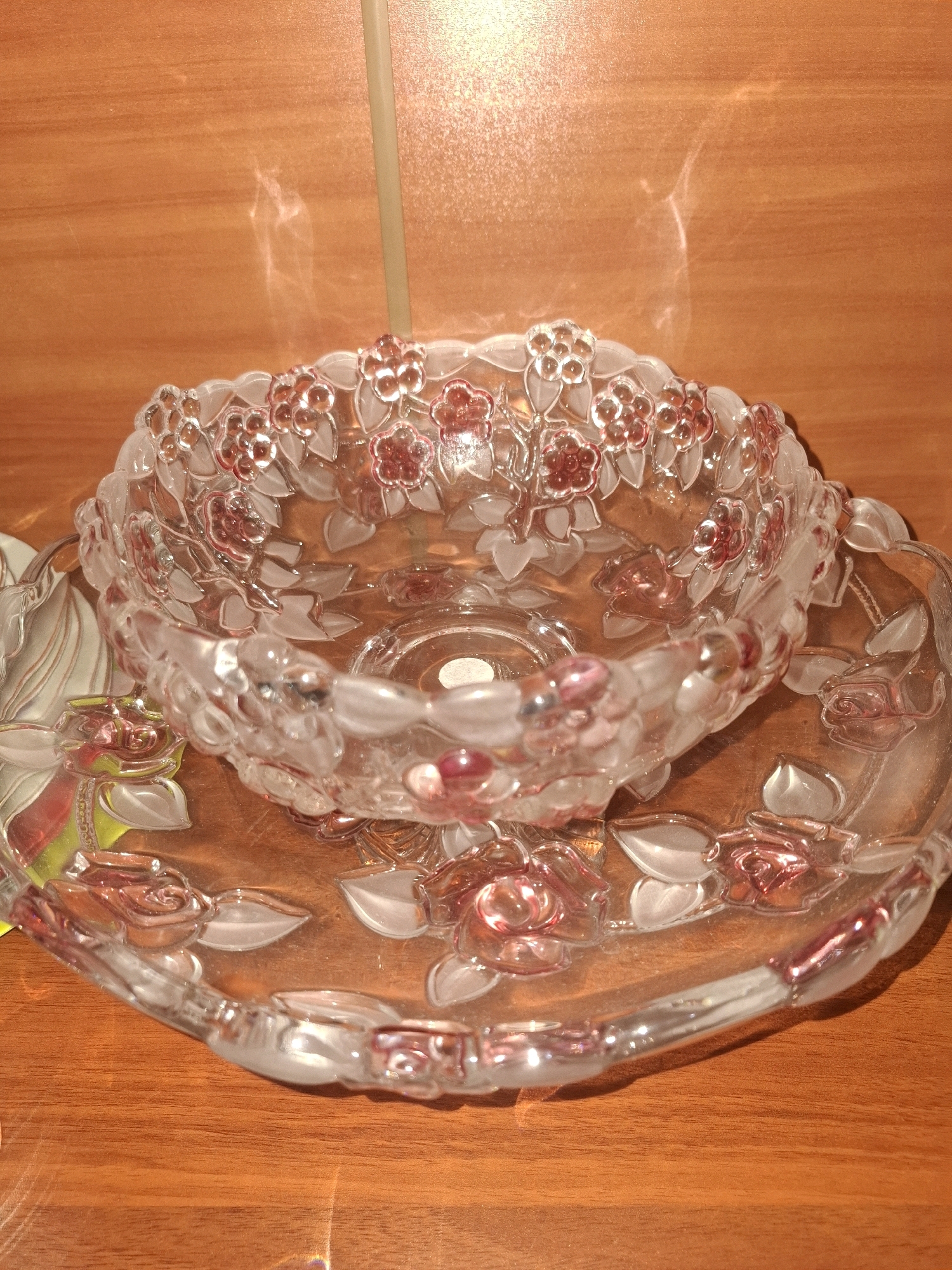 A decorative glass bowl with embossed floral designs, featuring pink and clear glass accents, is placed on a matching glass plate. The plate also has embossed floral designs, with a rose motif prominent. The items are positioned on a wooden surface, likely a table, which has a warm, orange-brown hue and a visible wood grain. The lighting in the scene is soft, creating reflections and highlights on the glass objects, and a vertical, pale yellow object is visible in the background, possibly a light fixture or a stick. There are no people present in the image. The location is the city of Almaty, Kazakhstan.