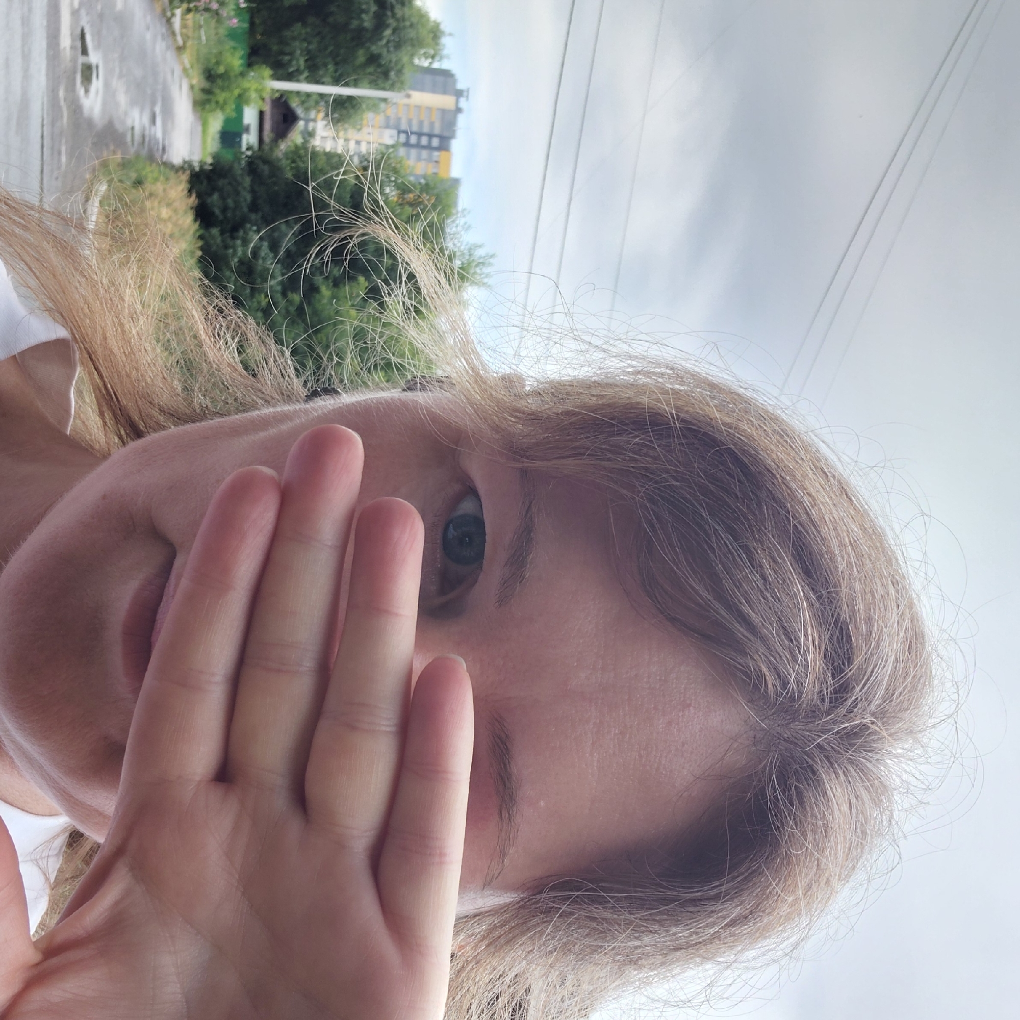 The image shows a close-up of a woman's face as she covers her left eye with her hand. She has light brown hair and is wearing a white shirt. Her right eye is visible, and it is green. The background is blurred, but appears to be an outdoor scene with trees and buildings. The sky is overcast. The location is identified as Novosibirsk, Russia.