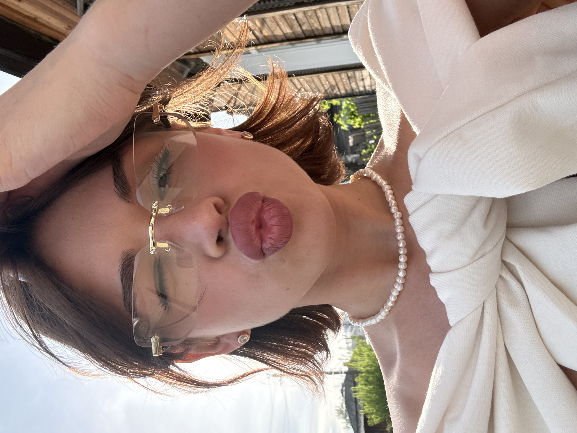 A woman is posing for a picture, tilted to the right. She has shoulder-length brown hair, fair skin, and is wearing gold-framed sunglasses. She also has a pearl necklace and a white garment tied with a knot. Her lips are prominent, pressed together and slightly pursed. Her hand is resting near her head.
In the background, some greenery, building, and sky can be seen. Considering the angle and the limited view, the specific location in Sarapul, Russia cannot be identified. The scene appears to be outdoors, possibly on a rooftop or a balcony.