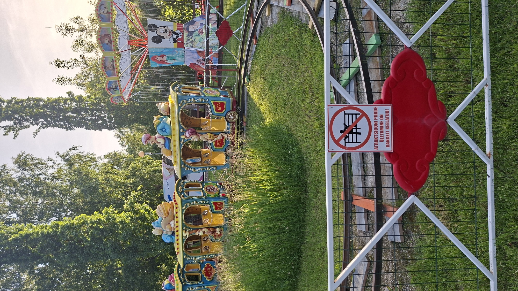 In Tallinn, Estonia, an amusement park scene unfolds. A colorful, multi-car train ride, adorned with cartoon characters, sits idle on its tracks amidst tall, green grass and trees. To the right, a swing ride, featuring images of Disney characters like Ariel and Mickey Mouse, is also stationary. In the foreground, a white metal fence with a red decorative element encloses the area. A sign on the fence displays a red circle with a diagonal line through it, indicating prohibition, and text in Estonian, "ATRAKTSIOONI PIIRDE ÜLETAMINE ON RANGELT KEELATUD!", which translates to "Crossing the attraction's boundary is strictly forbidden!". The sky is a soft, warm hue, suggesting either early morning or late afternoon. There are no people visible in the image.
