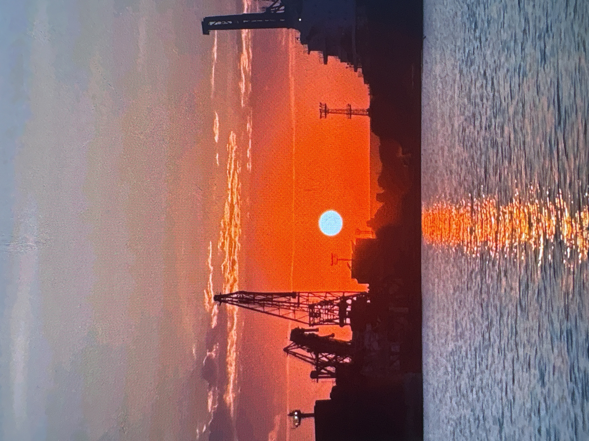 The image depicts a sunset over water, with a cityscape silhouette in the background. Cranes are visible in the foreground, suggesting a port or industrial area. The sun appears as a pale blue circle against an orange sky. The water reflects the orange color of the sunset. The location is described as Unknown, Unknown.