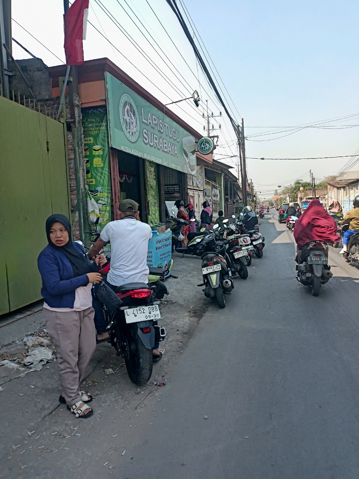 The image depicts a street scene in Surabaya, Indonesia. On the left side of the street, a woman wearing a dark hijab, a blue cardigan, and light-colored pants stands next to a parked motorcycle. She is looking to her left with an engaged expression. Behind her, a man wearing a white t-shirt and a green cap is seated on the motorcycle, also looking forward. The motorcycle has a license plate reading "L 4152 DBB 09-30". A storefront with a green facade and a banner displaying "LAPIS TUGU SURABAYA" is visible behind them. The street is paved and shows signs of wear. Further down the street, several other motorcycles are parked, and people are seen riding them. In the background, more buildings and utility poles with a web of electrical wires are present. The sky is clear and bright, indicating daylight.