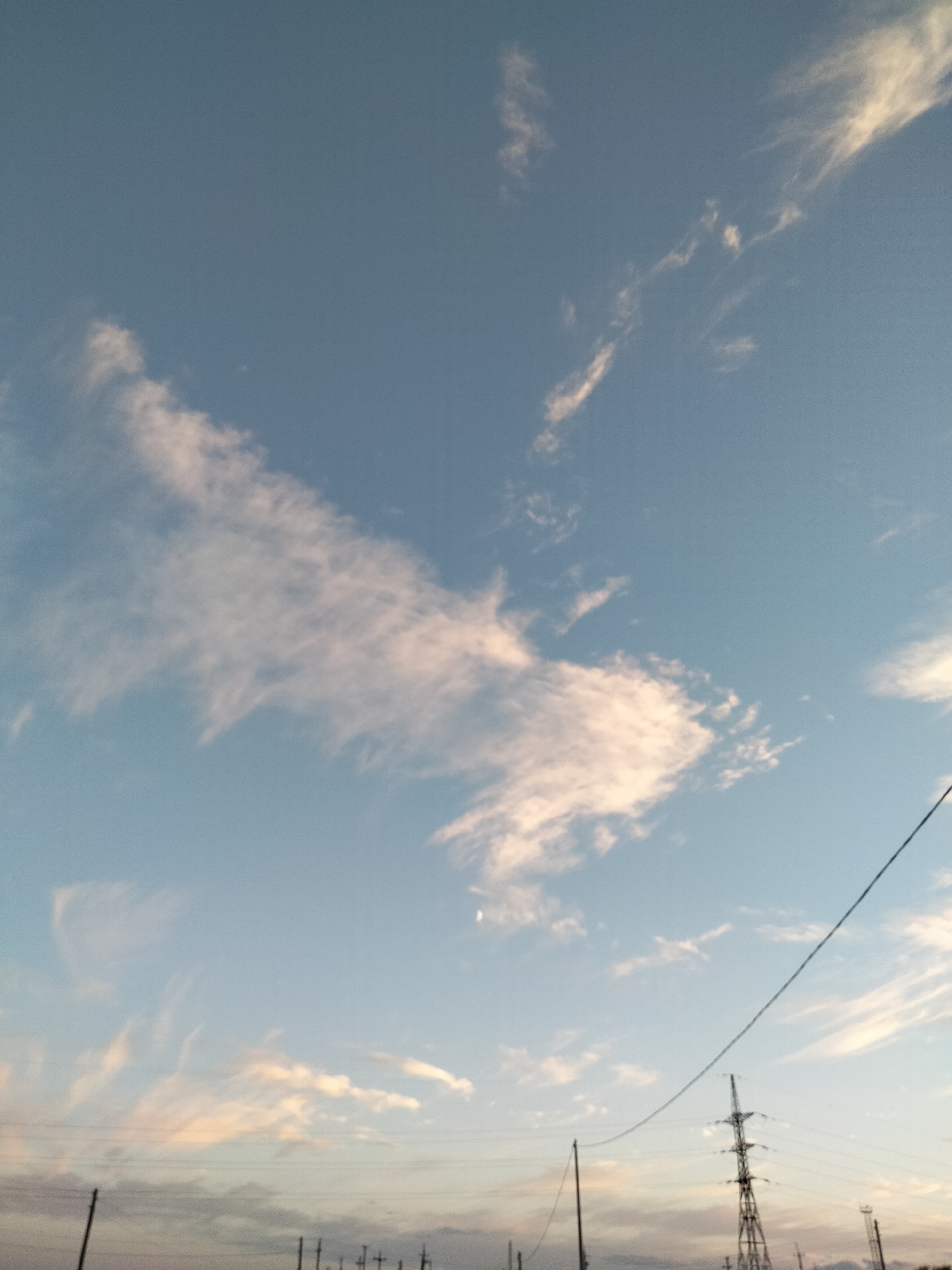 The image shows a wide-angle view of a sky with wispy clouds and a pale crescent moon visible. In the foreground, there are power lines stretching across the frame, supported by tall, lattice-style transmission towers. Some smaller poles are also visible along the horizon, along with what appears to be the silhouette of a distant urban landscape under a pale sky. The overall lighting suggests it may be either sunrise or sunset. The scene is devoid of any people or animals.