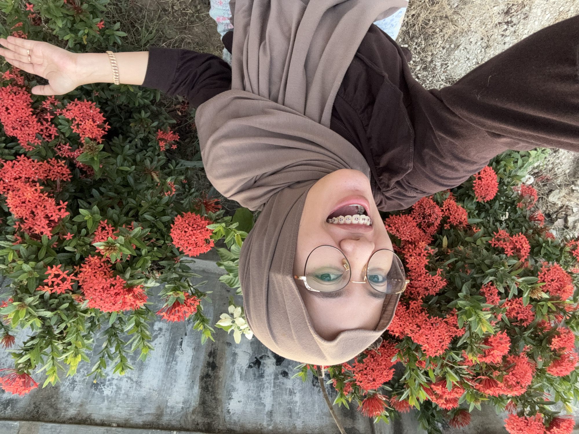 A young woman wearing a brown hijab and glasses is posing for a selfie outdoors in front of a wall and a bush with red flowers. She is smiling and has braces on her teeth. She is wearing a brown long-sleeved shirt and a bracelet on her left wrist. The background consists of a grey, textured wall and a dense bush with bright red flowers and green leaves. The ground is covered with small stones and dry grass. The image appears to have been taken during the daytime. The presence of the Ixora flowers (commonly known as Soka flowers in Indonesia) suggests a location with a tropical or subtropical climate, consistent with Gadingrejo, Indonesia.