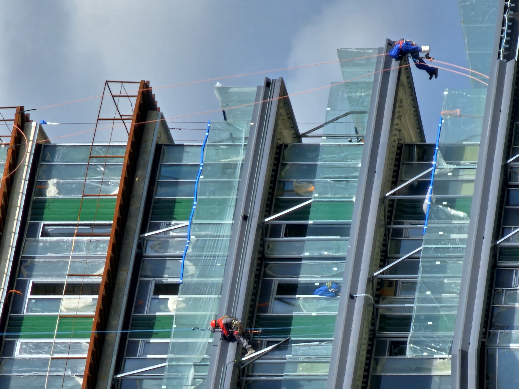 Two construction workers are rappelling down the side of a modern, glass-clad building under a cloudy sky. The building features a distinctive angular design with prominent vertical gray structural elements interspersed with glass panels, some of which are green. Orange safety lines are strung across the building facade, and blue ropes are also visible.
One worker, wearing a blue suit and a red harness, is positioned higher up on the structure, appearing to be actively working. The second worker, dressed in a red and black outfit with a helmet, is further down, also suspended by ropes and engaged in construction activities. The scene suggests ongoing construction or maintenance work on a skyscraper. The weather appears overcast, indicating it might be daytime, but the exact time is not discernible. There are no visible text elements in the image. The overall impression is one of an active construction site high above the ground.