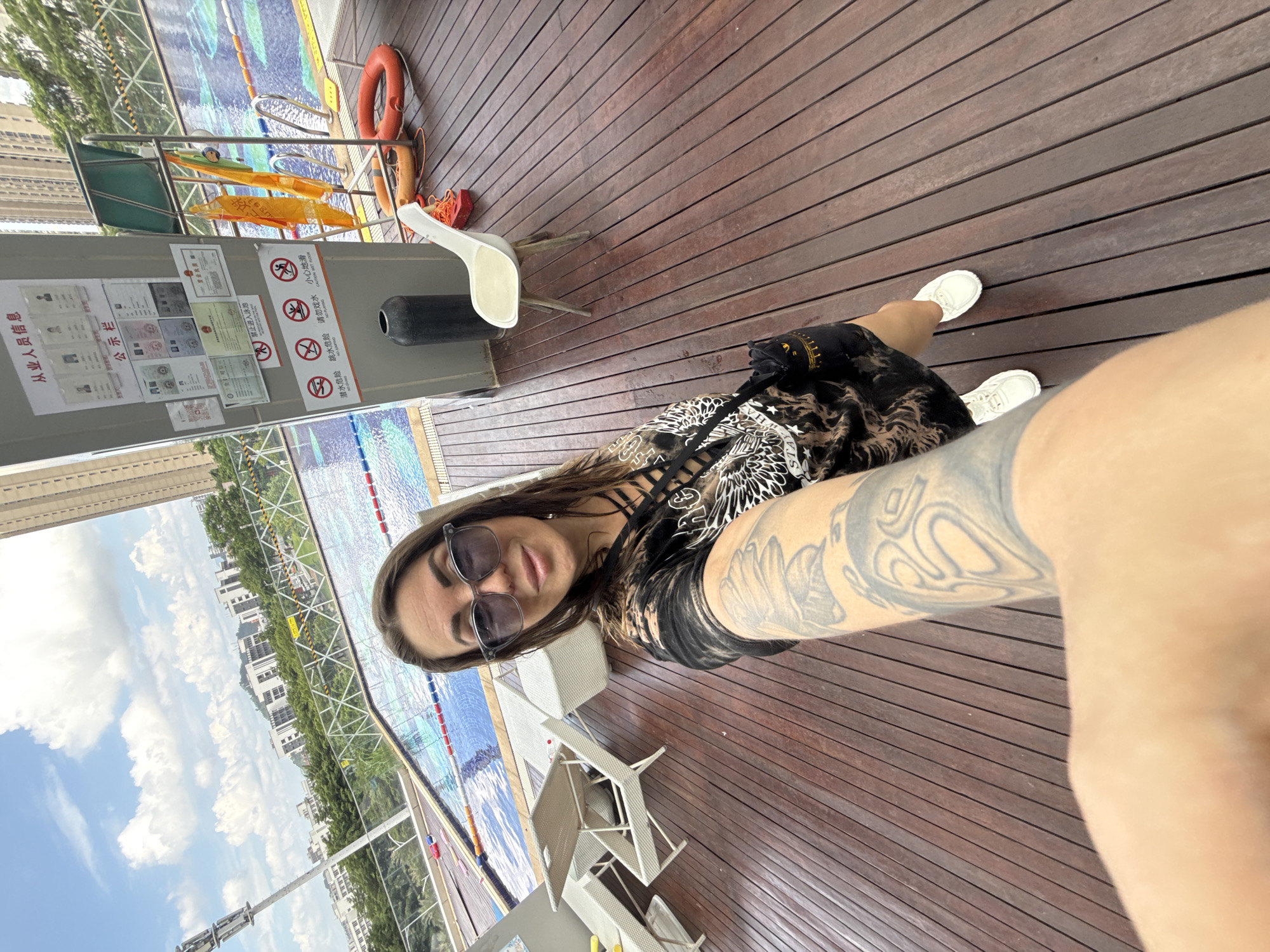 A woman is taking a selfie by a swimming pool in Huizhou, China. She has long, dark hair and is wearing sunglasses and a black t-shirt with a graphic print. She has tattoos on her arm. The swimming pool is blue with lane markers and is surrounded by a wooden deck. There are lounge chairs and a small table on the deck. In the background, there are tall buildings and lush greenery. A sign with Chinese text and several warning symbols related to pool safety is visible on a pillar next to the pool. A white chair and some safety equipment, including a lifebuoy, are also present. The sky is blue with scattered clouds.