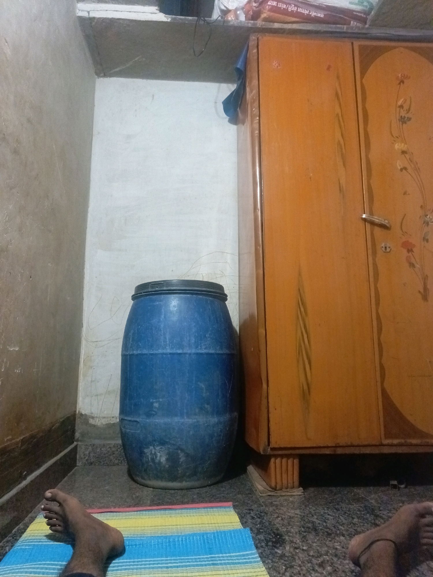 In a dimly lit room in Tippapura, India, a blue plastic barrel stands on a dark granite floor. The barrel is cylindrical with a black lid and appears to be full. To the right of the barrel, a tall, orange wooden wardrobe with ornate floral carvings on its doors dominates the scene. The wardrobe has a silver handle on its right door and appears to be elevated on wooden blocks. In the foreground, a colorful striped mat is laid out, with one bare foot with dark skin resting on it. The foot is positioned with the toes facing upwards, and a thin black anklet is visible around the ankle. The walls of the room are plastered, with the left wall appearing textured and a lighter color, while the back wall is white and shows some faint markings. A shelf is visible above the wardrobe, holding what appears to be a stack of books or packaged goods.