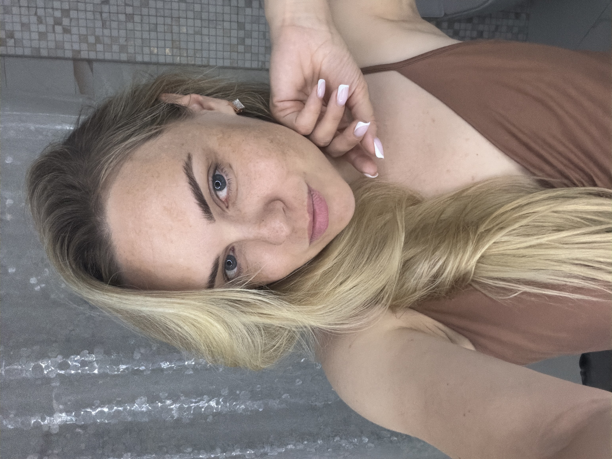 A woman is lying on a textured gray surface, possibly a tiled floor. Her head is tilted to the left, and she is looking directly at the camera. She has fair skin with freckles, blue eyes, and blonde hair styled to fall around her face and chest. Her left hand is near her face, showcasing a manicure with white tips. She is wearing a brown tank top. The background is a blurred tiled wall. The photograph appears to be taken indoors. There is no information to suggest this is taken in Votkinsk, Russia.