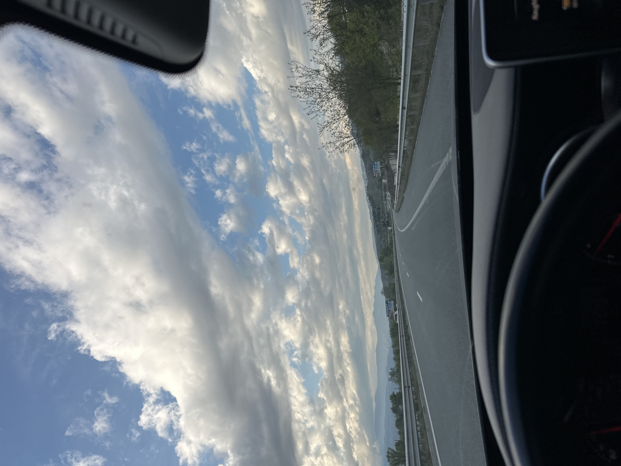 The image depicts a view from inside a car on a highway near Valkovo, Bulgaria. The sky is filled with clouds, some white and fluffy, with patches of blue sky visible. The highway stretches into the distance, flanked by trees and guardrails. A vehicle dashboard and steering wheel are visible in the foreground.