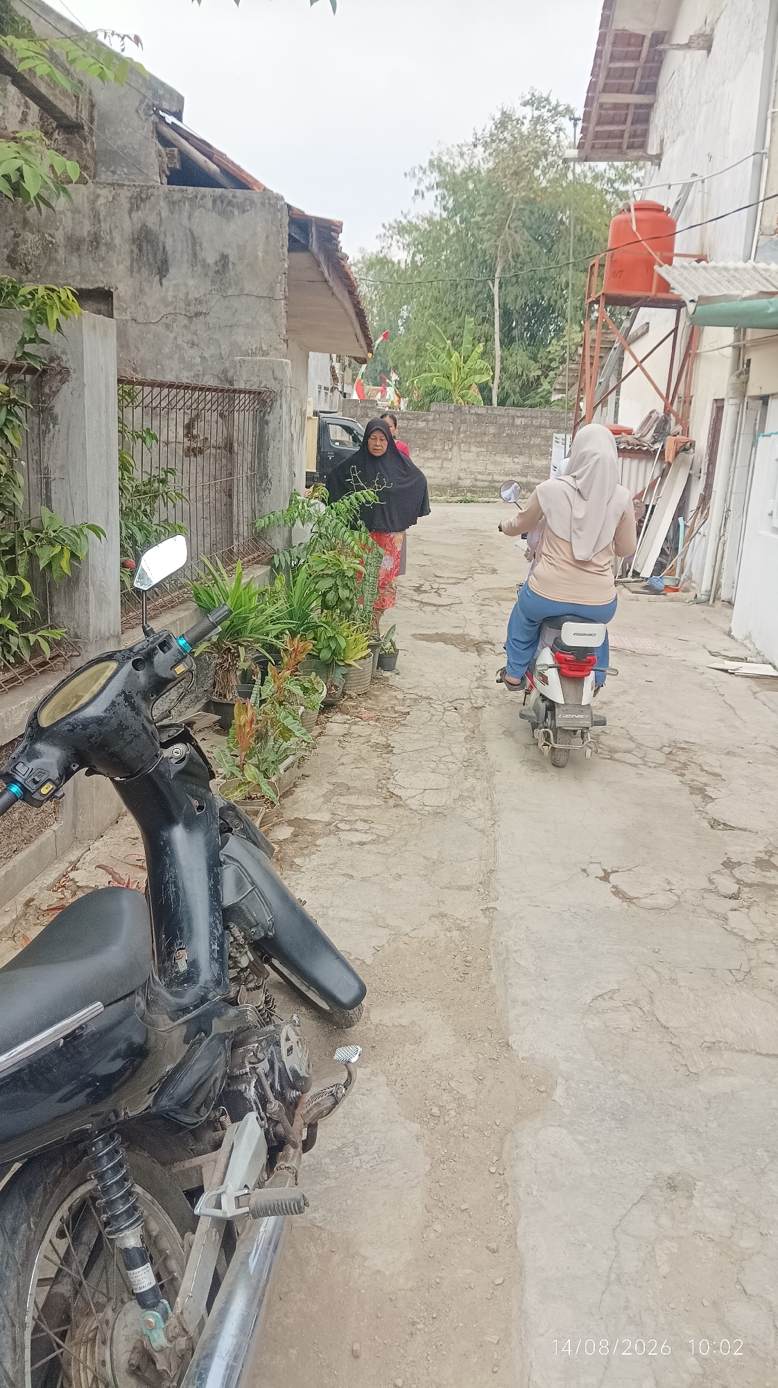 The image depicts an outdoor scene on a paved pathway, possibly a residential alleyway, in Tasikmalaya, Indonesia. In the foreground, a black motorcycle is parked, angled towards the left. Further down the pathway, a woman wearing a light brown hijab and blue pants rides a white and red scooter away from the viewer. To the left of the pathway, a woman in a black hijab and a patterned skirt stands next to a black car and a row of potted plants. The buildings visible in the background appear to be simple houses with concrete walls and tiled roofs. An orange water tank is mounted on a metal structure on the right side of the image. The ground is a mix of cracked concrete and dirt. The sky is overcast. The timestamp "14/08/2026 10:02" is visible in the lower right corner.