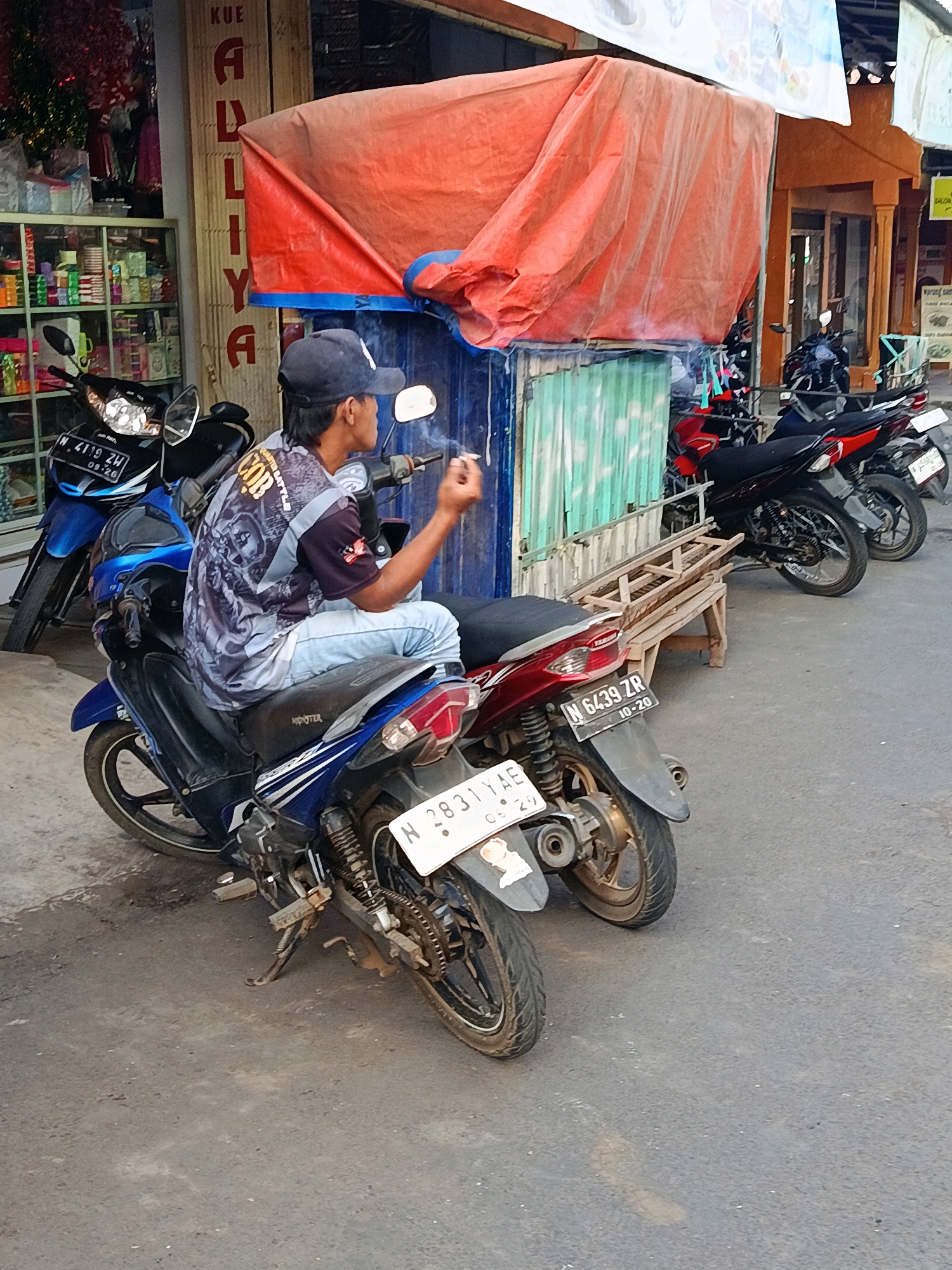 The image shows a street scene in Senduro, Indonesia. In the foreground, a man wearing a dark blue baseball cap and a patterned t-shirt is sitting on a blue and black motorcycle. He is holding a cigarette in his right hand and appears to be smoking. The motorcycle has a license plate that reads "N 2831 YAE". To the left of this motorcycle is another blue motorcycle with a license plate reading "N 4119 ZW". Behind the man, there are several other motorcycles parked, including red and black ones. A wooden bench is positioned near the motorcycles. In the background, there is a building with a red tarp draped over part of its facade, and shelves stocked with items are visible through a storefront window to the left. The ground is paved with asphalt.