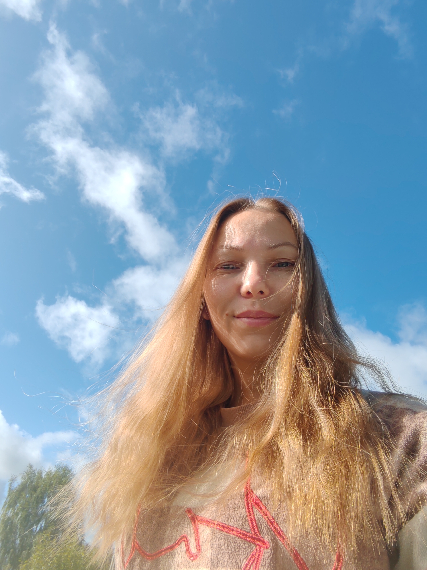 A selfie captured outdoors shows a young woman with long, blonde hair against a bright blue sky with scattered white clouds. She is looking directly at the camera with a slight smile. Her hair is windswept, with some strands falling across her face. She is wearing a light-colored, possibly beige, sweater with a red embroidered pattern across the chest. The lower left corner of the image shows the top of green foliage, suggesting the presence of trees or bushes. The lighting indicates it is daytime, likely sunny. There is no discernible urban environment or specific location markers in the frame.