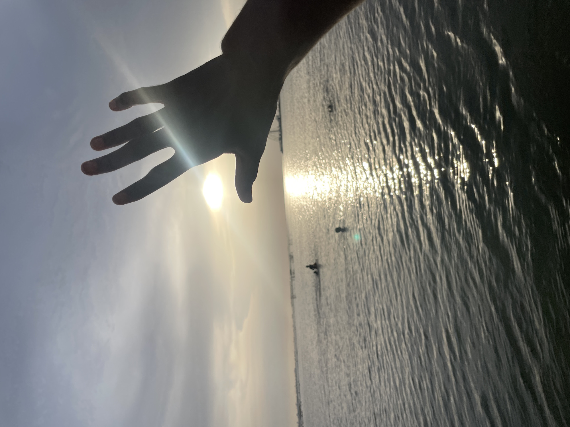 A serene, outdoor scene depicts a silhouetted human hand stretching from the upper left towards the center-right of the frame. The hand is open with fingers slightly spread, creating a strong silhouette against the bright sun positioned just below the palm. Intense lens flares emanate from the sun.
The background features a vast body of water, likely a lagoon or lake, which fills the lower half of the image. The sun's light reflects powerfully off the water's rippled surface, creating shimmering, elongated streaks of light that stretch towards the horizon. In the distance on the water, several small, dark, and indistinct shapes are visible, appearing as silhouettes against the bright reflections. These shapes could be distant boats, people, or large birds.
Above the water, the sky transitions from a soft, hazy light blue on the left to a brighter, near-white hue closer to the sun. The sky is partly cloudy but generally clear, contributing to the overall bright and atmospheric feel. The strong backlighting and low sun angle indicate either a sunrise or sunset. The image evokes a sense of peace and connection with nature, highlighting the interplay of light and shadow over a tranquil body of water. The setting is in Ikorodu, Nigeria. No text is visible in the media file.