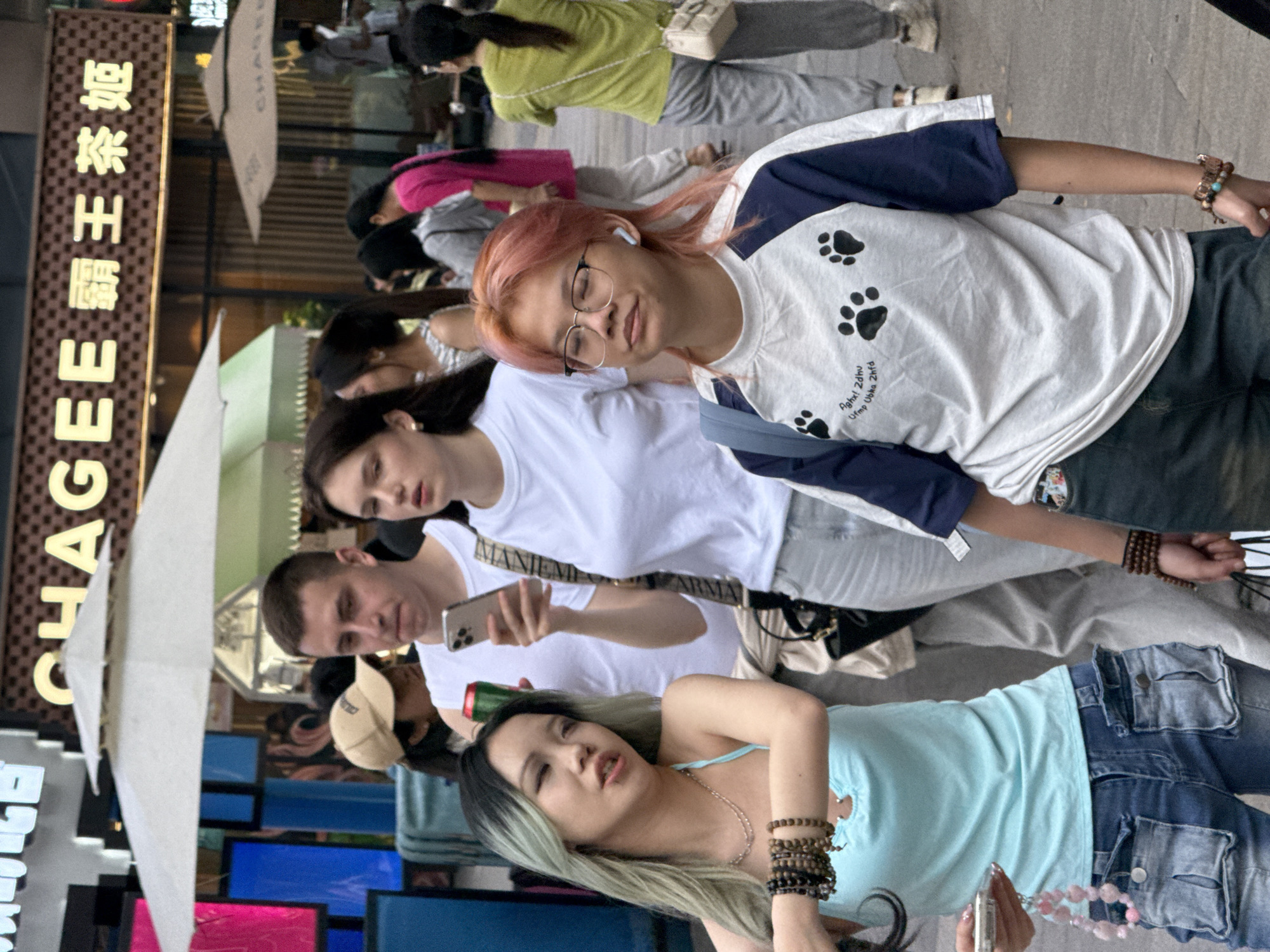 The image captures a group of individuals standing on a city street, likely in the Yuzhong District of China. The presence of Chinese characters on a vertical sign reading "CHAGEE 霸王茶姬" suggests this location.
The individuals are dressed in casual clothing and appear to be tourists or locals exploring the area. At least one person is holding a smartphone.
The setting includes a combination of modern structures and street furniture, with umbrellas providing shade. The scene suggests a bustling urban area with a blend of commercial and social activity.