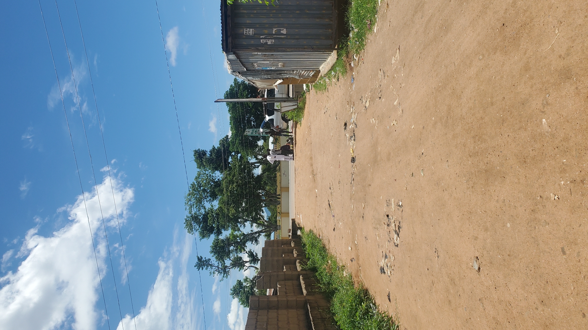 The image depicts a dirt road in Jalingo, Nigeria. The road is unpaved and shows signs of wear and tear, with visible debris scattered across its surface. On the left side of the road, a brick wall extends into the distance, and on the right, a corrugated metal structure, likely a shop or container, is present. In the background, several people are gathered near a white vehicle with its trunk open. Trees line the background, with a bright blue sky and white clouds above. The scene appears to be a typical street view in a developing urban area.