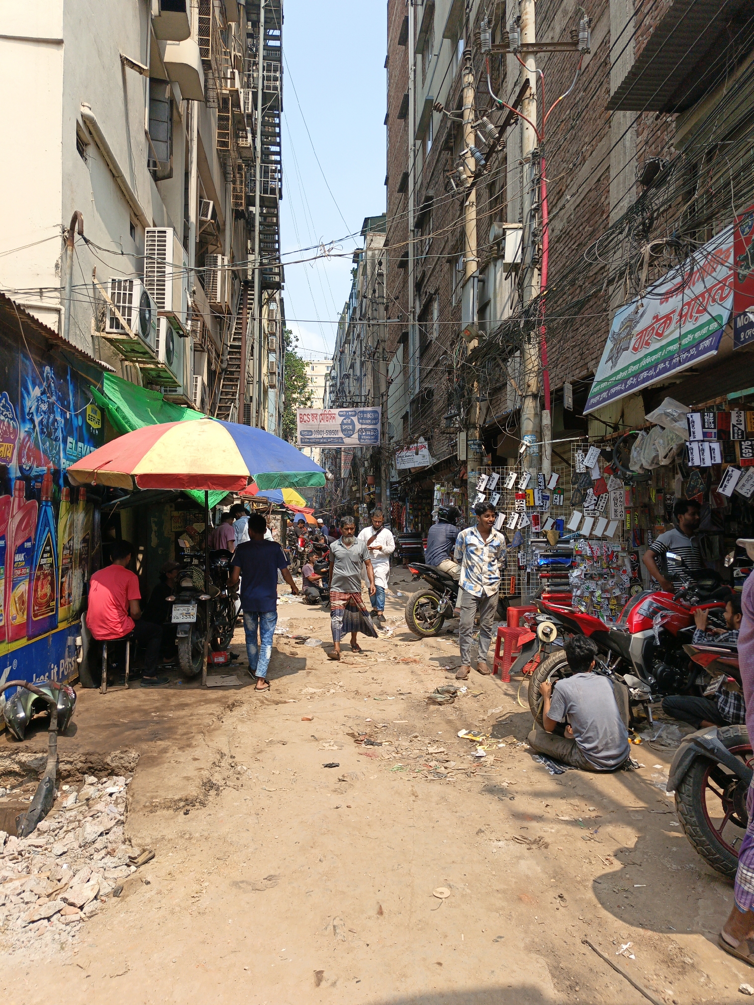 This image depicts a narrow, bustling street in Dhaka, Bangladesh. The ground is unpaved and covered in dirt and debris. People are walking and gathered around various shops and stalls. There are motorcycles parked and being worked on. The buildings are tall and closely packed together, with electrical wires crisscrossing overhead. The shops display signs in Bengali. One shop has a large blue and red advertisement for engine oil. There are colorful umbrellas providing shade for vendors. The general atmosphere is one of activity and commerce.