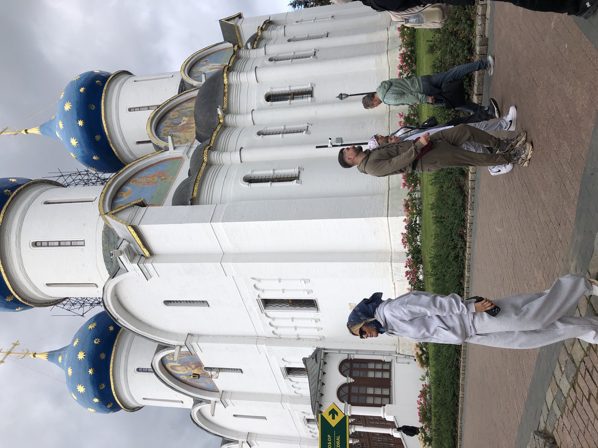 In an outdoor scene, a white, multi-domed church with blue domes adorned with gold stars dominates the background. Several people are visible walking on a paved path in front of the church. In the foreground, a woman wearing a grey tracksuit and a blue headscarf is walking towards the left of the frame, holding a phone in her right hand. To her right, a group of three people are walking together. A man in a dark olive tracksuit is in the lead, followed by a child in a white top and a woman with a patterned headscarf. Further to the right, another man in a light green jacket and blue jeans is walking with a black bag. The ground is paved with reddish-brown bricks, and a strip of greenery with pink flowers runs along the base of the church wall. A yellow sign with an arrow points to the left, with text that appears to be in Russian. The sky is overcast.