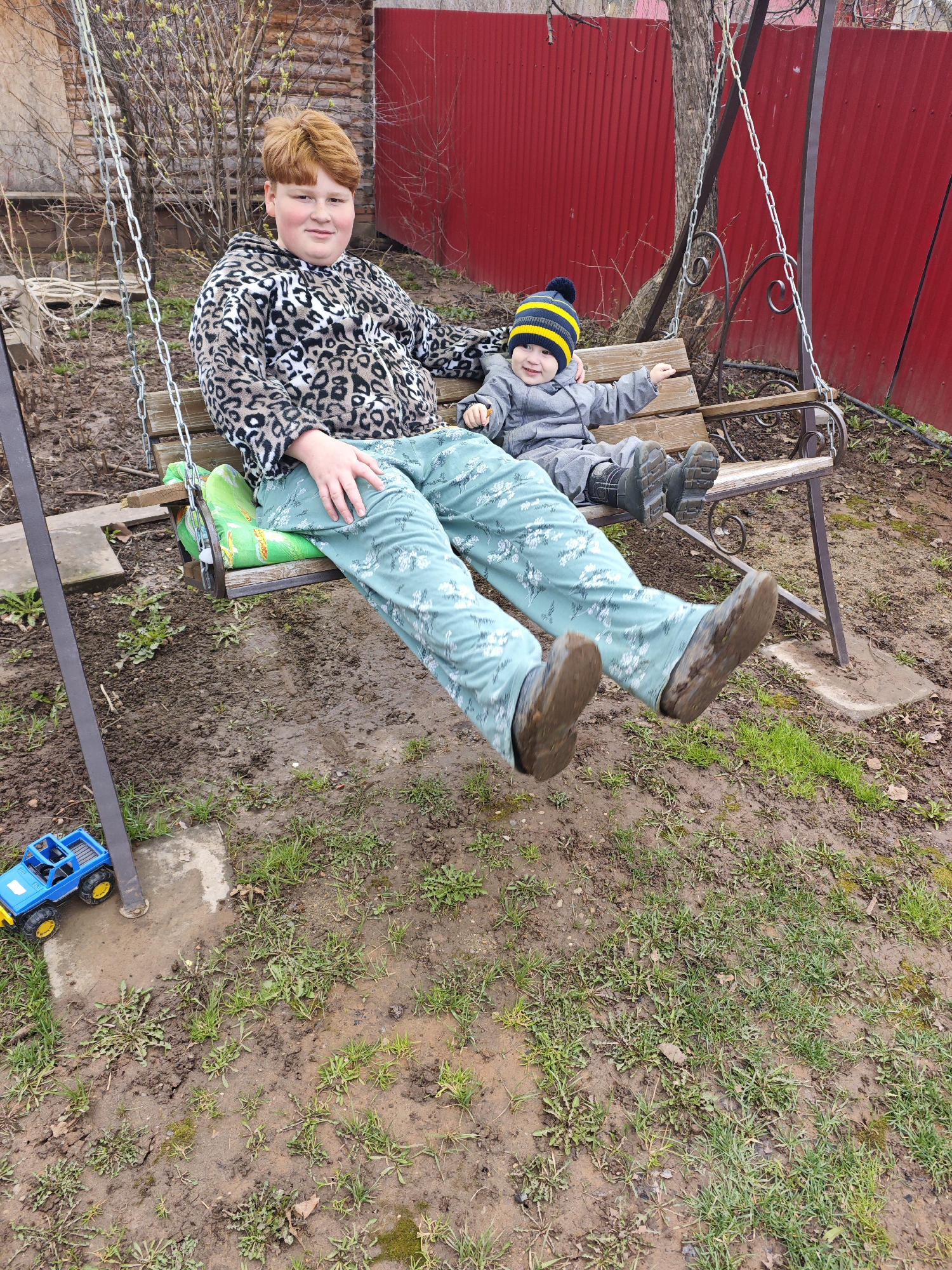 This vertical shot captures two boys on a garden swing in a rural or backyard setting in Foki, Russia, likely on an overcast day given the diffused light. The ground beneath the swing is muddy with sparse patches of green grass and puddles, suggesting recent rain.
On the left, an older boy with reddish-brown hair and a light complexion sits wearing a leopard print hooded jacket and light green patterned trousers. He looks towards the camera with a slight smile. His feet, clad in dirty boots, are suspended mid-air. Beside him, on the right, a younger boy, likely a toddler, is seated. He wears a grey padded jumpsuit and a blue and yellow striped knitted beanie with a pom-pom. The younger boy smiles broadly, looking slightly to his right, with his arm outstretched. His small boots are also covered in mud.
The swing features a wooden slatted seat with ornate dark metal armrests and is suspended by metal chains from a dark metal frame. A bright green cushion is visible on the swing seat. In the background, a striking red corrugated metal fence stretches across the scene, with a bare tree showing early buds peeking over it. To the left, a rustic wooden structure, possibly a shed or part of a building, is visible. A blue toy truck with yellow wheels rests on the muddy ground near the swing's frame on the left side of the image. There is no visible text in the image.