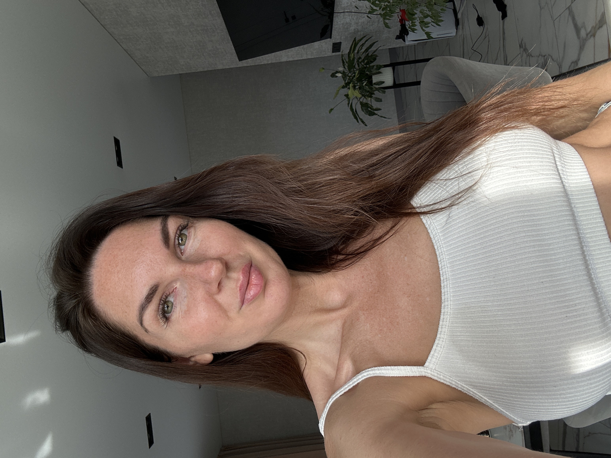 The image is a close-up selfie of a woman with long, brown hair and green eyes. She is wearing a white ribbed tank top. Her skin has a light complexion with freckles. The background appears to be an indoor setting, possibly a modern apartment, with a gray textured wall, a large black television mounted on the wall, and a potted plant. The lighting suggests it is daytime, with sunlight casting shadows on the ceiling. The flooring is a marble-like tile. There is no indication of the city of Chaikovsky, Russia, in the image.