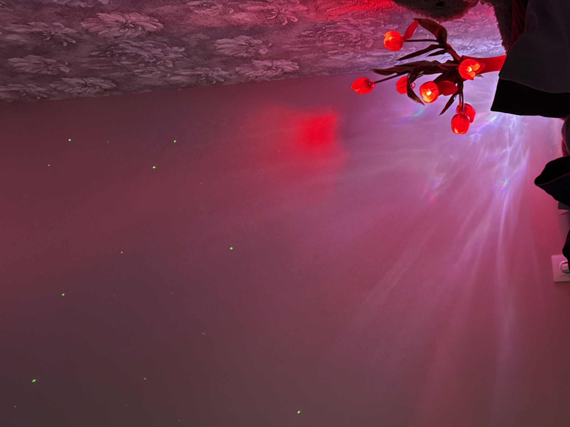 The image captures an indoor scene dominated by ambient lighting and decorative elements. The upper portion reveals a ceiling covered in what appears to be floral-patterned wallpaper. Below this, a plain wall is illuminated with a soft, pervasive red and pink glow. Scattered across this wall are multiple small, bright green dots, suggesting the use of a laser projector.
In the upper right corner, a decorative light fixture shaped like a branch adorned with several red, tulip-shaped lights is visible, with some of the tulips emitting a warm, soft light. These lights create shimmering, wave-like patterns and colorful reflections on the wall and ceiling, hinting at a dynamic light source like a disco ball or water effect projector. A dark-colored object with a white or light-colored trim, possibly a piece of clothing or a hanging fabric item, is partially visible in the top right corner, partially obscuring the ceiling and some lights. Towards the bottom right, a light-colored electrical socket or switch is partially visible on the wall. The overall lighting suggests an evening or nighttime setting, creating a cozy or atmospheric mood. No individuals are present in the image, and no text is discernible.