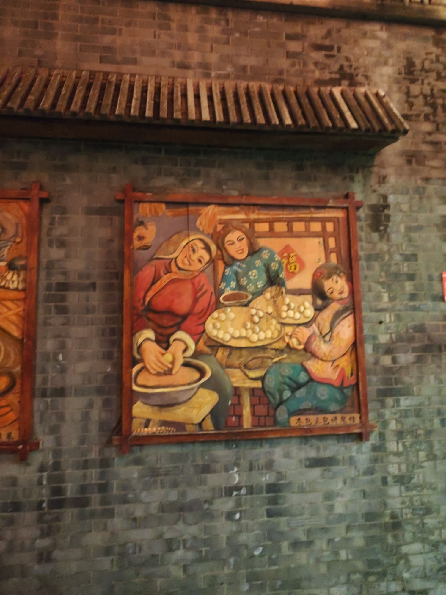 This image shows a colorful bas-relief carving mounted on a weathered brick wall. The carving depicts a traditional Chinese scene with three women engaged in what appears to be food preparation. The woman on the left, wearing a red and white outfit, is kneading dough in a large bowl. To her right, a woman in a green and white patterned garment is looking towards a plate piled high with round, light-colored objects, possibly dumplings or buns. The third woman, on the far right, is wearing a pink and yellow outfit and is also working with dough, with a contented expression. The carving is framed by a dark wooden border. The brick wall behind the carving is rough and grey, with visible mortar lines. A wooden roof structure with tiled accents runs above the carving. The overall impression is of a cultural artifact displayed outdoors in a historical or traditional setting. While no specific markings in the image directly indicate the location in Lindian, China, the style of the artwork and the architecture are consistent with traditional Chinese cultural displays.