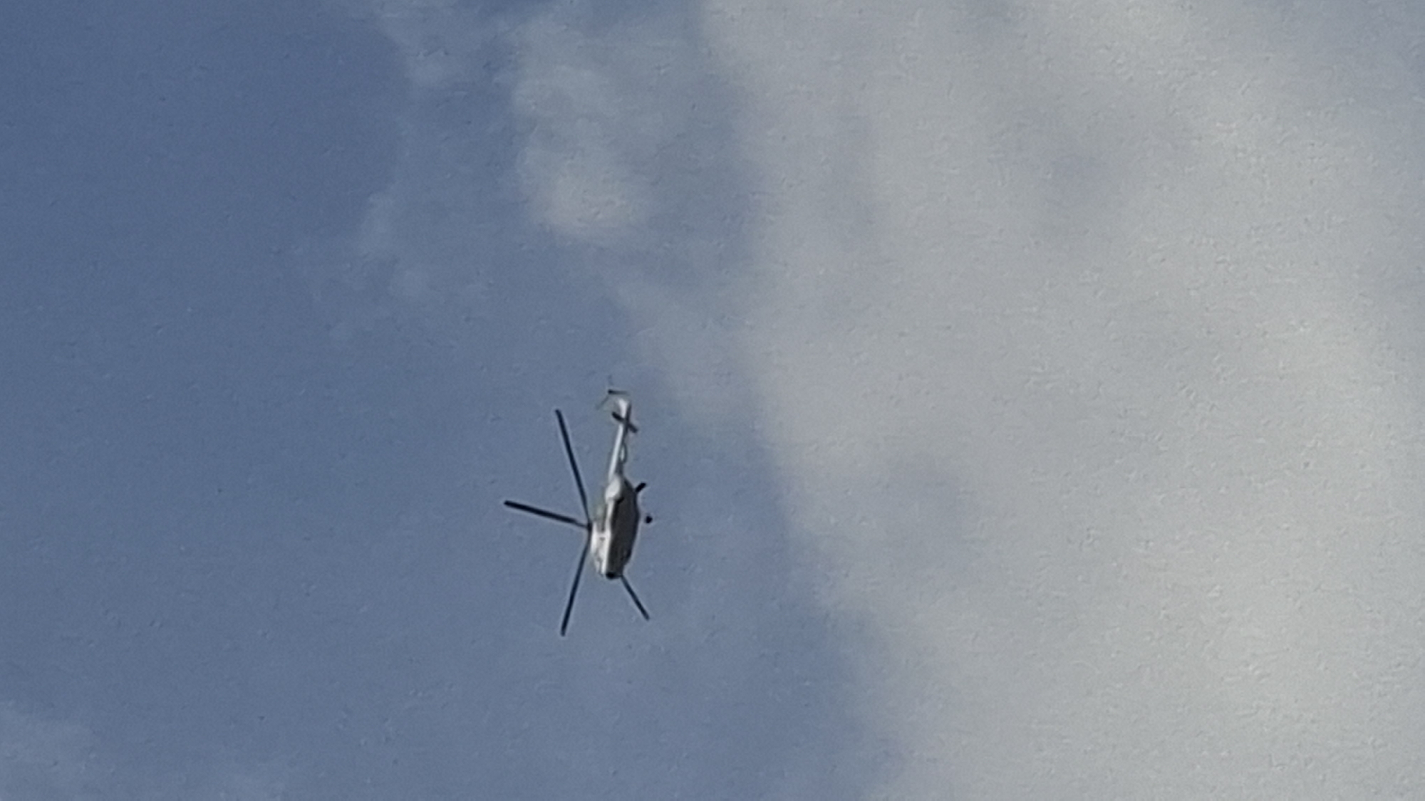 A helicopter is captured from below as it flies through a cloudy, blue sky. The helicopter is white and has a silver tail rotor. Its main rotor blades are blurred, indicating movement. The helicopter appears to be the sole subject of the image, with no people or other objects visible. The sky is partially obscured by large, white clouds, suggesting it could be a partly cloudy day. The lighting indicates it is daytime, but the specific time is not discernible. There are no clear location cues, and the image conveys no discernible emotion. No text is visible in the media file.