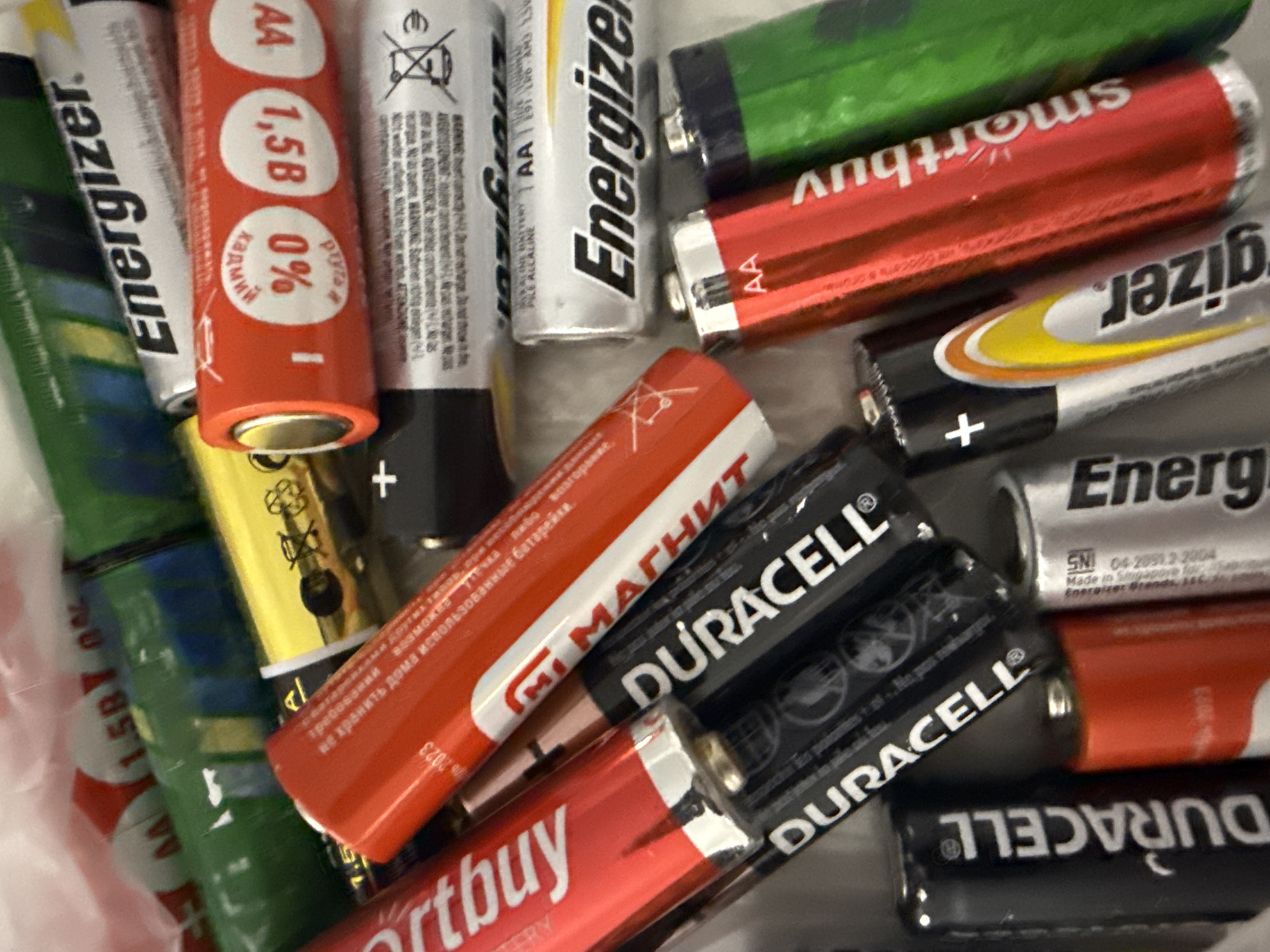 A close-up shot shows a collection of used batteries of various brands and types, likely discarded or awaiting recycling. Brands include Energizer, Duracell, Smartbuy, and other batteries labeled in Russian (such as "Магнит" (Magnet) and "1.5B"). Most appear to be AA size. The batteries are in a jumbled pile, with some showing signs of wear and tear. The location is uncertain; the image does not confirm the location is Votkinsk, Russia. The presence of Russian-labeled batteries suggests a Russian-speaking country.