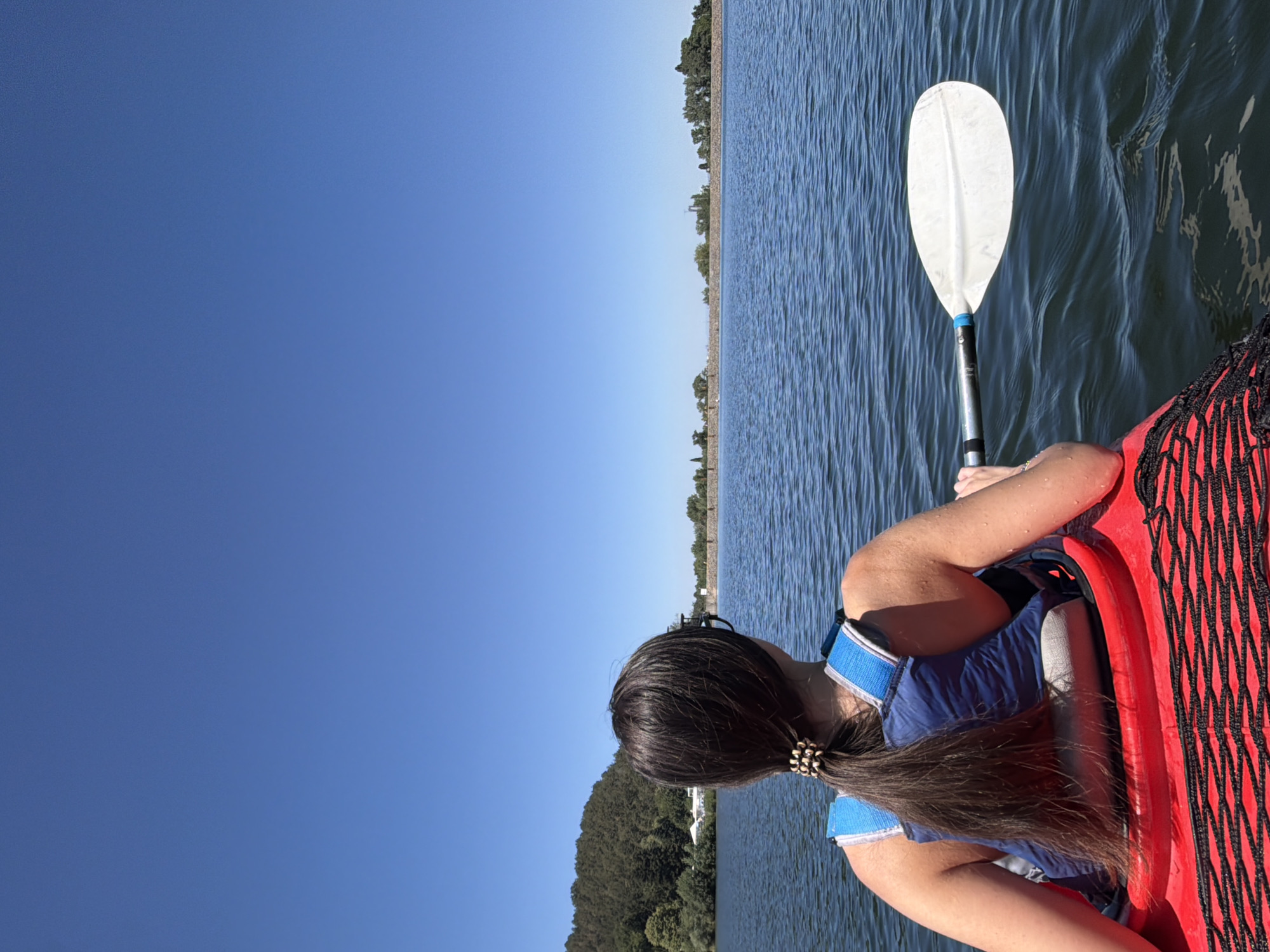 A woman with dark hair pulled back in a ponytail is seated in a red kayak, viewed from behind. She is wearing a blue life vest. Her right arm is extended, holding a white kayak paddle, which is resting on the surface of the water. The kayak is on a calm body of water, likely Lake Pancharevo in Pancharevo, Bulgaria, given the context. In the background, a tree-covered hill is visible on the left, and a long, stone embankment with trees and some structures stretches across the horizon on the right. The sky is clear and bright blue. The water has small ripples on its surface.