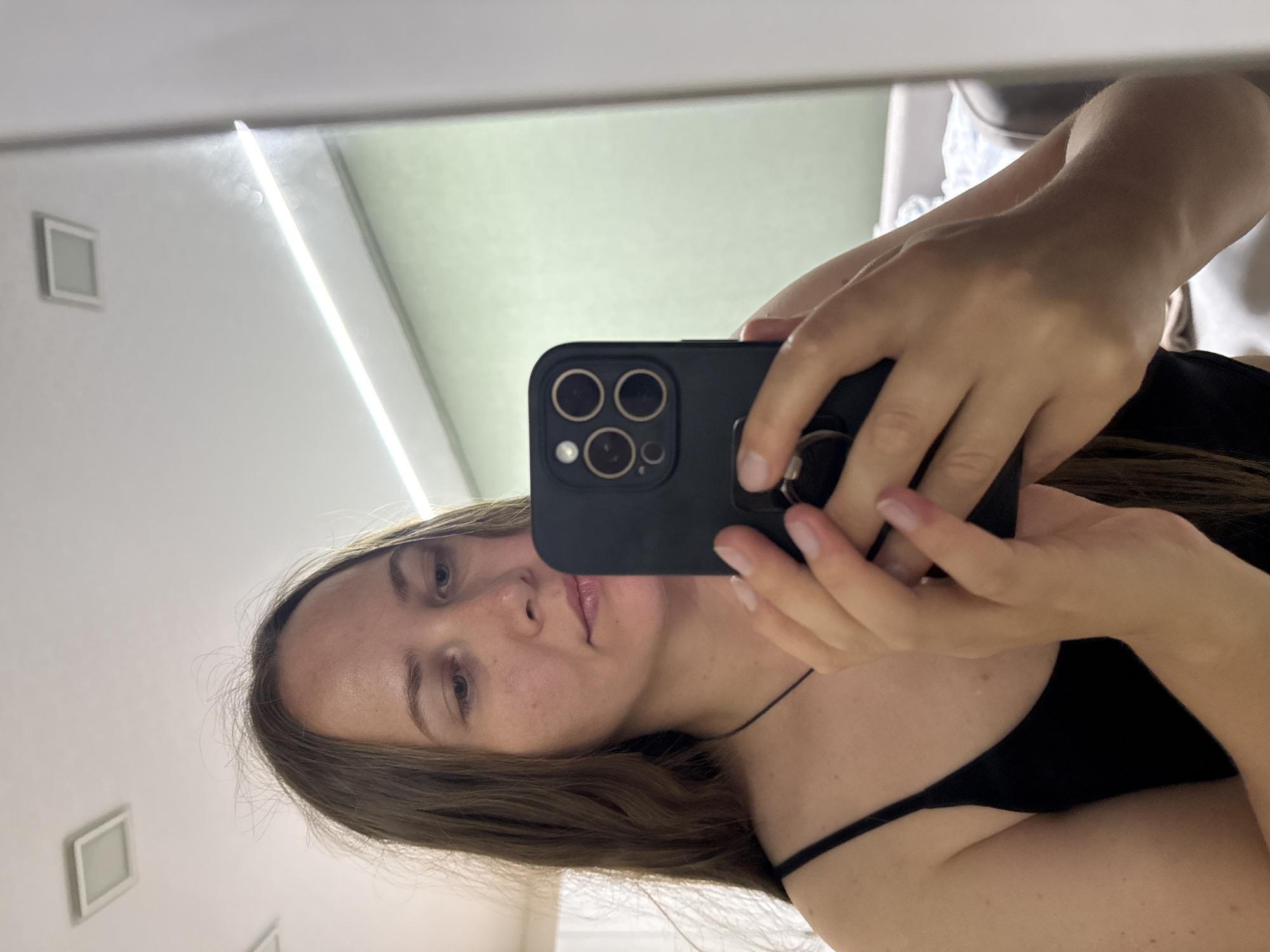 A young woman with long, brown hair is taking a mirror selfie indoors. She is wearing a black tank top and holding a black smartphone in her right hand, with her left hand supporting the phone. The phone case has a distinctive quad-camera design. The background appears to be a room with a white ceiling that has recessed lighting fixtures and a long, thin light strip. A light green wall is visible behind her. There is a hint of furniture and fabric in the blurred background to the right, suggesting it may be a bedroom or living area. The image is taken from a frontal perspective, focusing on the woman and her phone.