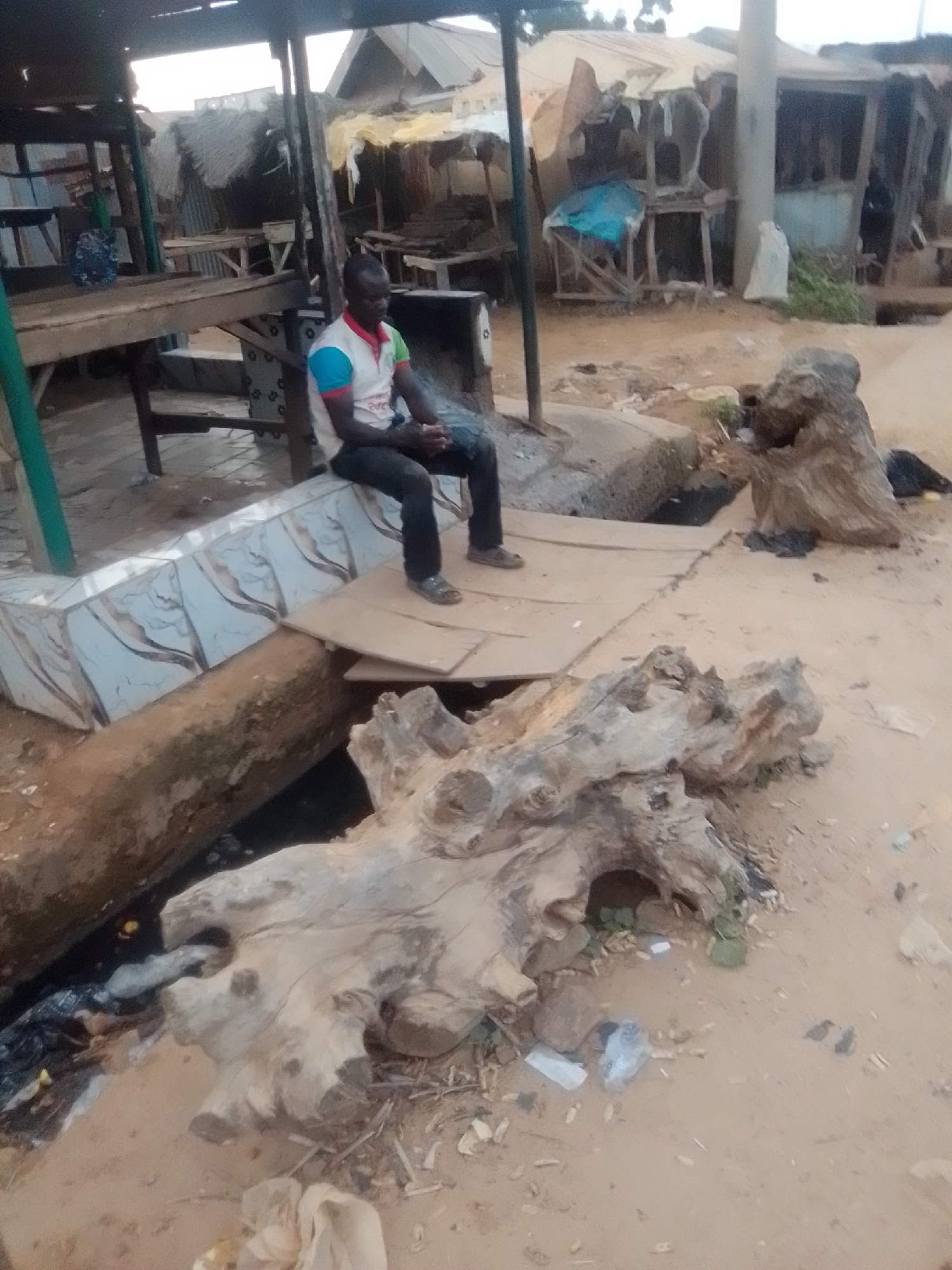 A man sits on a makeshift wooden platform at an outdoor market in Gombe, Nigeria. The man, wearing a colorful shirt and dark pants, is positioned in the center of the image, looking to his right. The market appears to be in a state of disrepair, with various stalls constructed from corrugated metal and wood. Debris, including plastic bags and other refuse, is scattered on the ground, which is covered in sand. A large, weathered tree trunk lies on the ground in the foreground, adding to the rustic and somewhat unkempt atmosphere of the scene. The sky is overcast, suggesting a cloudy day.