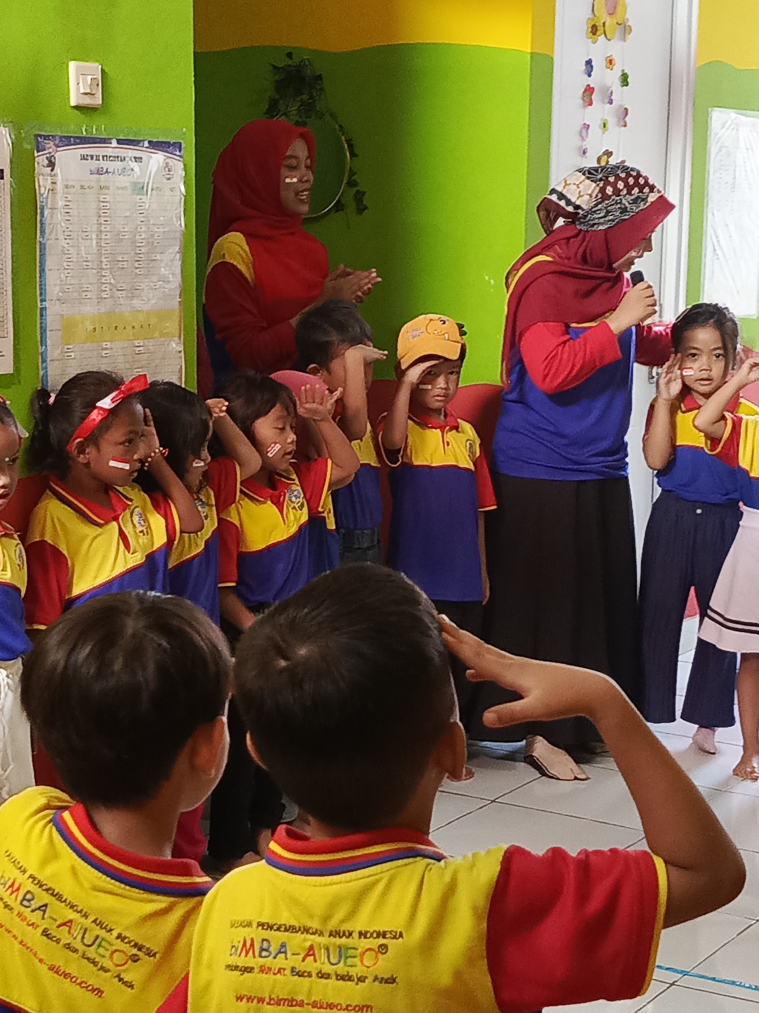 In Ciseeng, Indonesia, a group of young children, dressed in yellow and red shirts, stand in formation while some salute, their faces adorned with red and white face paint. Two adults, one a woman in a red hijab and the other partially obscured, stand facing the children. The room features a bright green wall with a light switch and a bulletin board displaying schedules and announcements. Decorations, possibly flowers, hang from the ceiling. The floor is tiled. The overall scene depicts an educational or patriotic event for children.