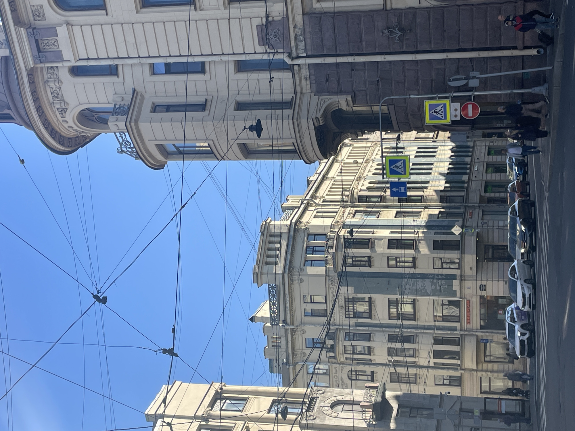 The image depicts a street scene in Saint Petersburg, Russia, on a clear, sunny day. The architecture is characteristic of the city, with multi-story buildings featuring ornate detailing, mansard roofs, and large windows. Trolley wires crisscross the sky, indicating the presence of public transportation. Several cars are visible on the street, suggesting moderate traffic. Pedestrians are walking on the sidewalks and crossing the street at a designated crosswalk. Traffic signs, including pedestrian crossing signals and a "no entry" sign, are posted. The buildings are in various states of repair, with some showing signs of wear. The overall impression is of a bustling urban environment with a rich historical backdrop.