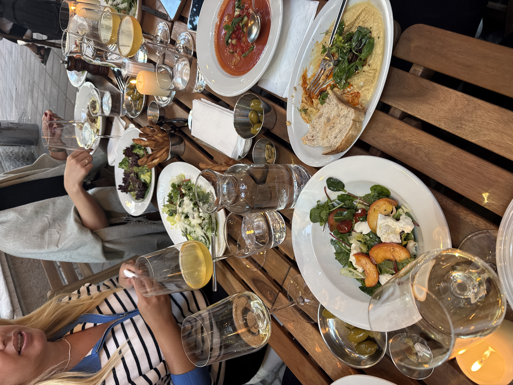 The image depicts an outdoor dining scene at a restaurant in Belgrade, Serbia. A wooden table is laden with various dishes and drinks, suggesting a meal in progress. In the foreground, two wine glasses filled with white wine are prominently displayed, one tilting slightly. A salad with peaches and cheese sits in a white bowl to the right of the wine glasses, and a plate of hummus with bread and greens is visible to the right of that. Further back, a bowl of gazpacho soup with garnishes is on the table, along with a silver container holding green olives and small dishes of what appear to be capers or seeds. A bread basket with grissini sticks is also present. Several water glasses and a carafe of water are on the table. Two lit candles add a warm ambiance. In the background, two women are partially visible, seated at the table. One woman, in a striped shirt, is holding a white napkin. The other woman, wearing a light grey shawl, has her hands on the table. The setting appears to be a pleasant evening meal.