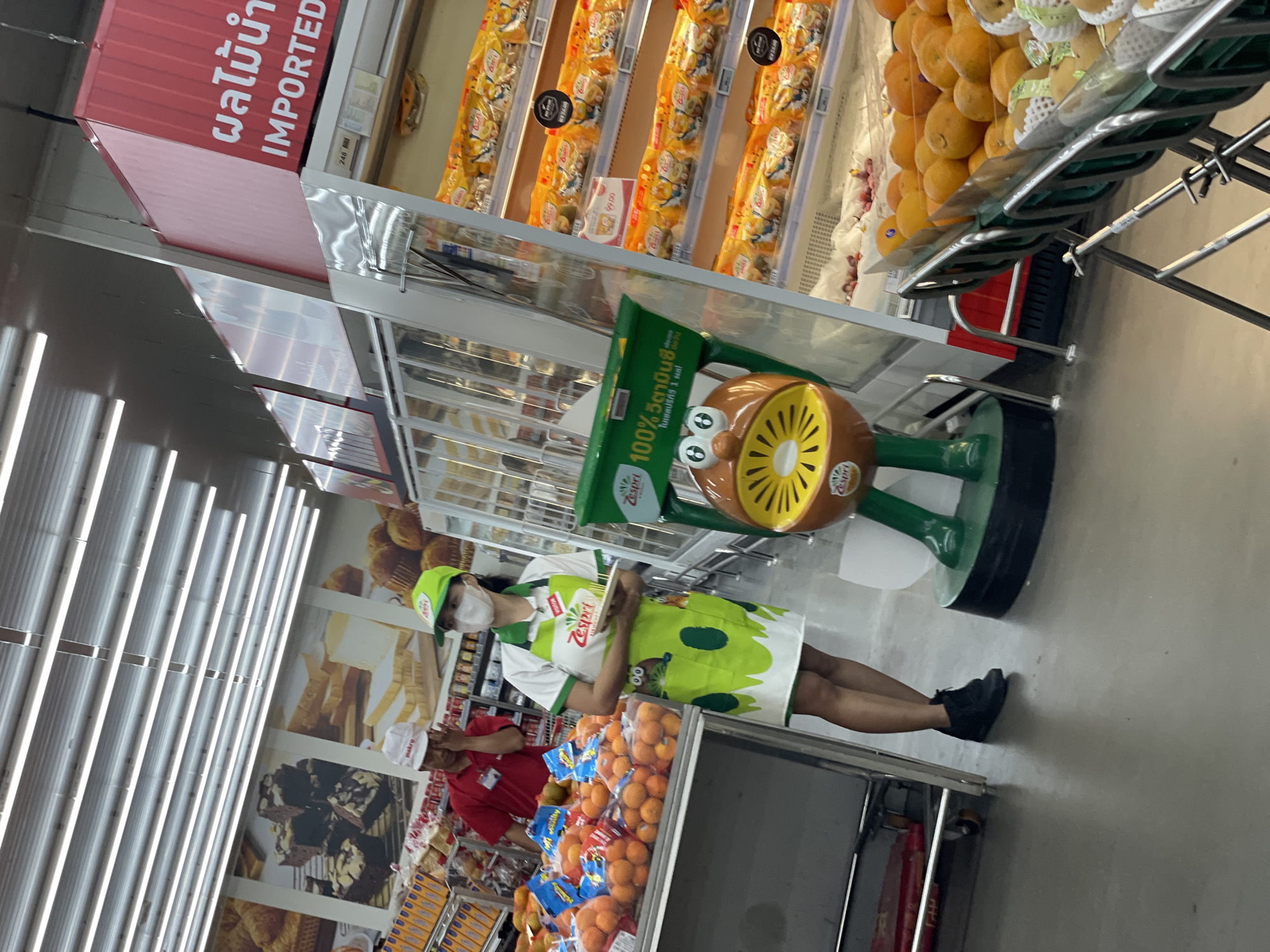 The image shows the interior of a supermarket, likely a Makro store in Pattaya City, Thailand. In the foreground, a woman wearing a green Zespri apron and a white face mask stands to the right of a display of oranges. She is holding a small white plate with some items on it. To her left is a large, cartoonish kiwi fruit stand. Further back, a man wearing a red shirt and a white cap is visible, appearing to be an employee. The background shows shelves stocked with packaged goods, some featuring bright yellow packaging. The lighting appears to be fluorescent, typical of a retail environment. The overall scene depicts a retail setting with a focus on produce and packaged food items.