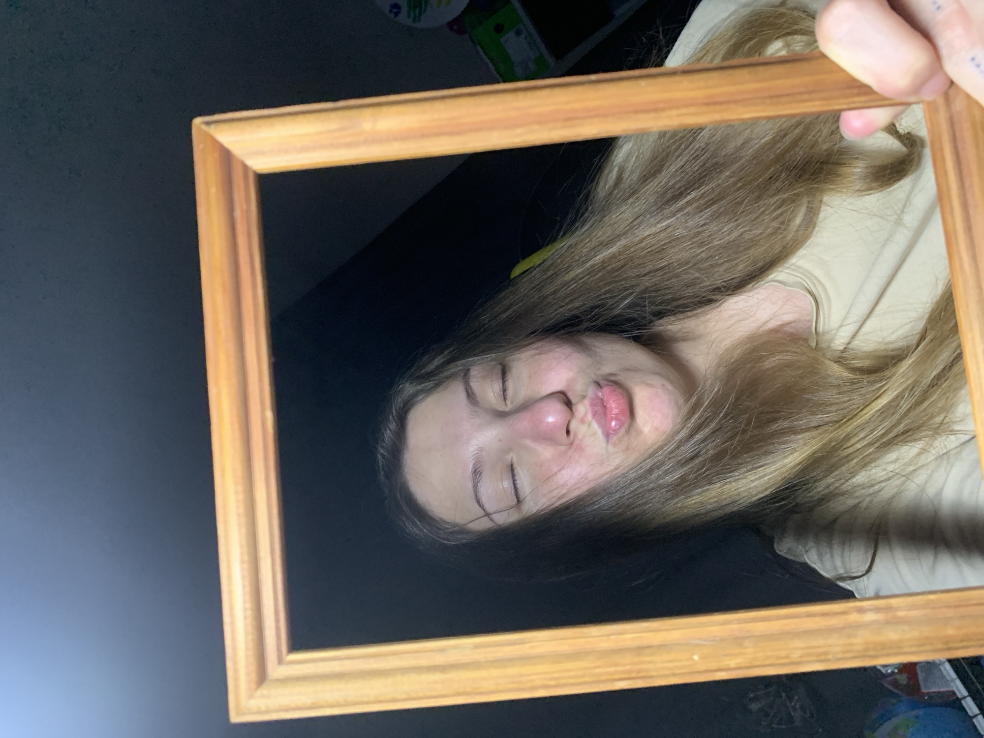 A young Caucasian female with long, light-brown hair is shown in a close-up shot, looking directly at the camera with her eyes closed and lips pursed in a prominent puckering or kissing gesture. She holds a simple, light-brown wooden picture frame vertically in front of her face, framing it from approximately her forehead to her chin. Her hands are visible at the bottom of the frame, gripping the sides. The subject's complexion shows a subtle reddish tint, particularly around the nose and cheeks. She wears a plain, light-colored (likely beige or tan) crew-neck top. The overall scene is dimly lit indoors, with the background appearing predominantly dark. Faintly discernible objects in the periphery include what appears to be a blue globe or spherical item on a surface to the lower left, and various items, including a green box and a multi-colored disc, on shelves to the right, indicating a domestic environment.