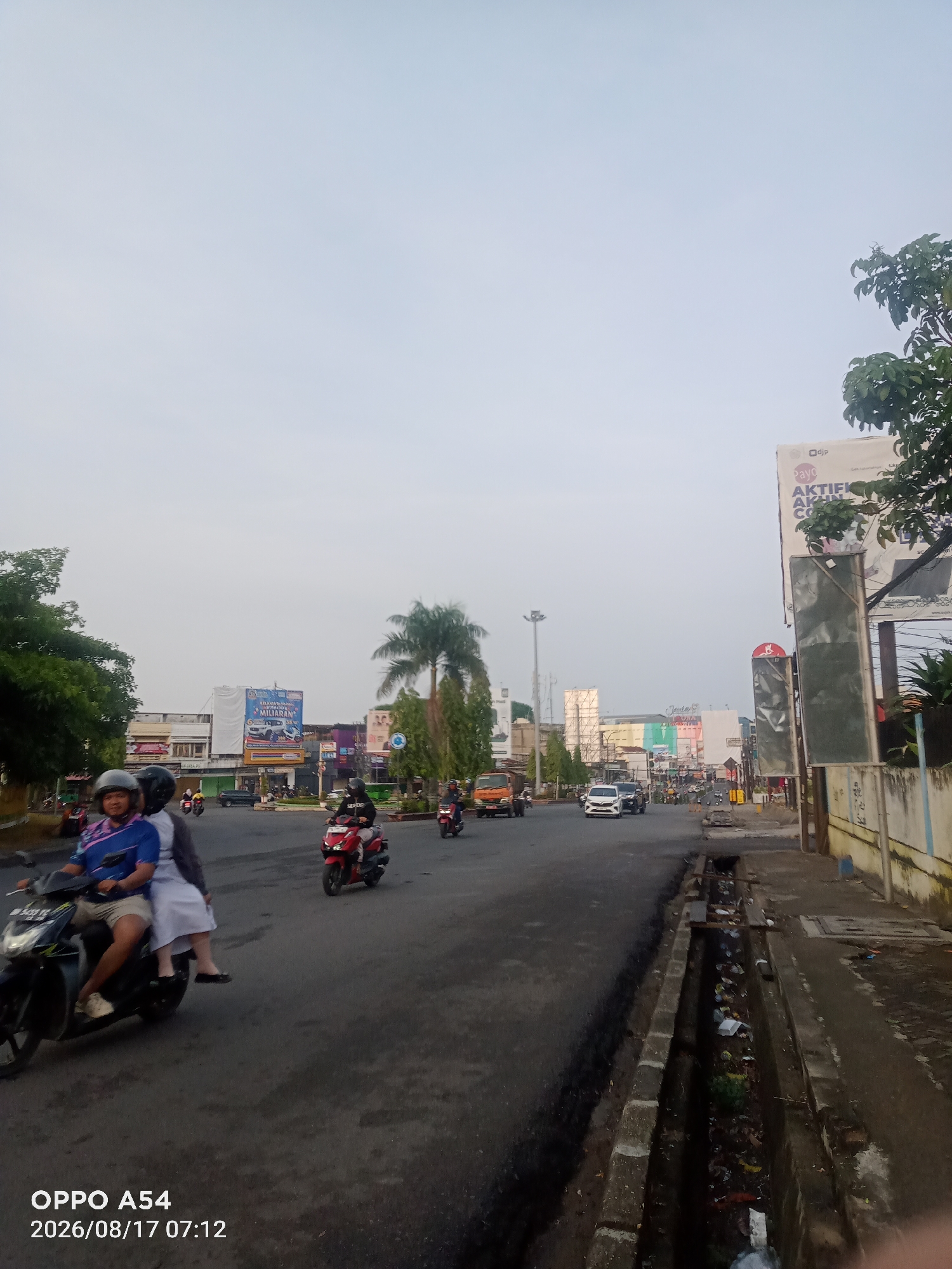 The image depicts a street scene in Jambi, Indonesia, with a focus on a road and its surroundings. In the foreground, a dark asphalt road is visible, with a concrete drainage ditch running along the right side. The ditch is filled with debris, including plastic bags and other trash. Several motorcycles are present on the road. One motorcycle in the left foreground carries two people, the rider wearing a blue shirt and a helmet, and the passenger wearing a white outfit and a dark scarf. Further down the road, other motorcycles are seen, some with riders and some without clear occupants. In the background, there are various buildings and billboards. One prominent billboard features text and an image of a car, likely an advertisement. Trees and palm trees are scattered throughout the scene, some lining the road and others appearing in courtyards. The sky is overcast and light blue, suggesting it might be early morning or late afternoon. The overall impression is of a busy urban street with traffic and commercial activity.