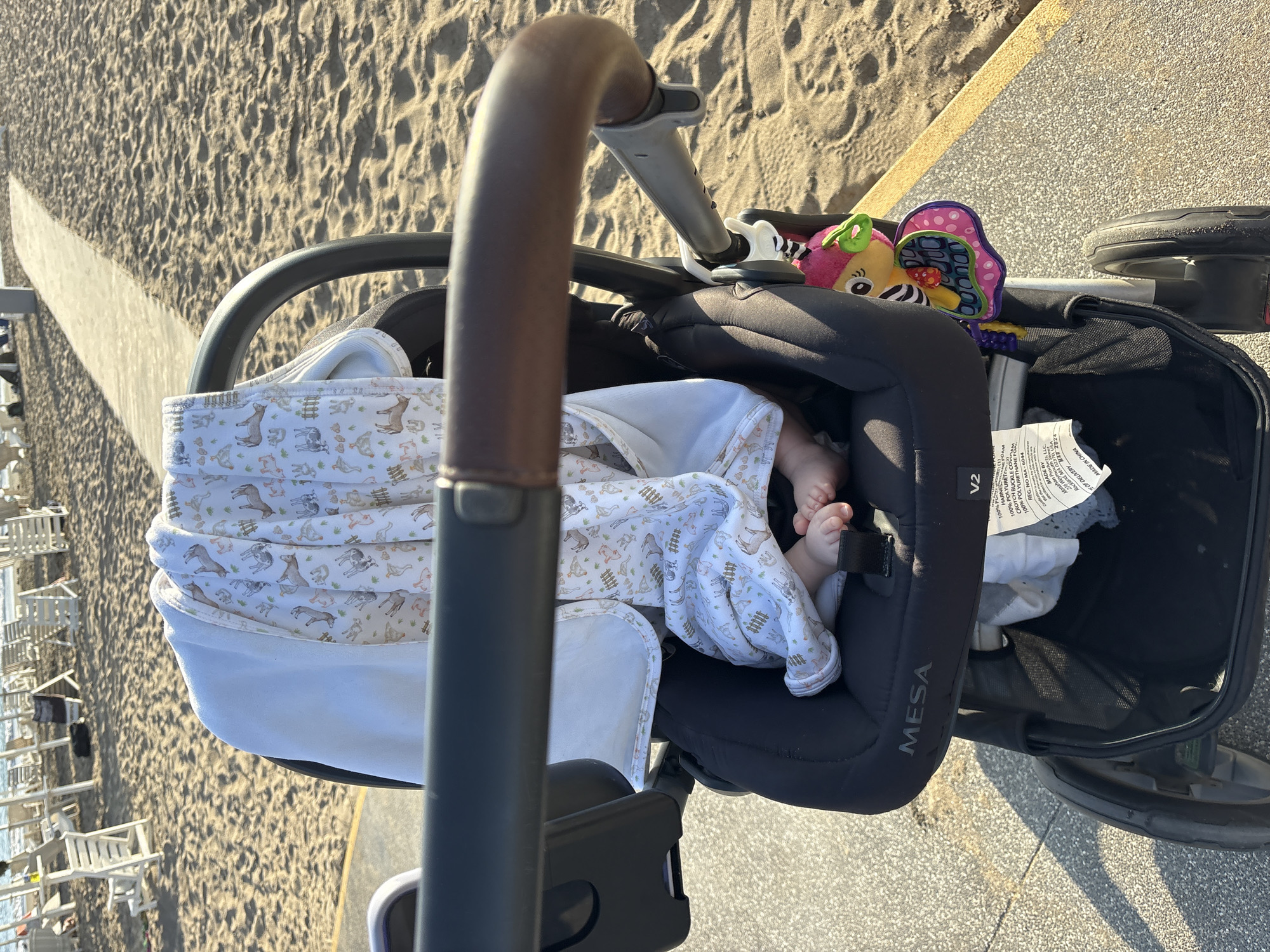 The image shows a baby stroller on a sandy beach pathway. The stroller is black with a brown handlebar. A baby's feet, covered by a blanket with a pattern of farm animals, are visible inside the stroller. A colorful toy is attached to the stroller. In the background, beach chairs and the ocean are visible. The scene is lit by sunlight, indicating it is daytime. The location is identified as Chorrillos, Peru.