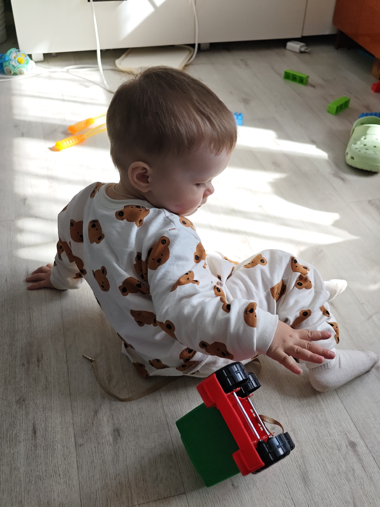 A young child wearing a white onesie with brown bear prints is seated on a light-colored, wood-grain floor. The child is looking down and touching a toy truck that is red with black wheels and a green block attached. Various toys are scattered on the floor, including a green clog, orange jump ropes, and green and blue building blocks. The scene is well-lit, possibly from sunlight entering the room. The location is in the city of Уральское, Russia.